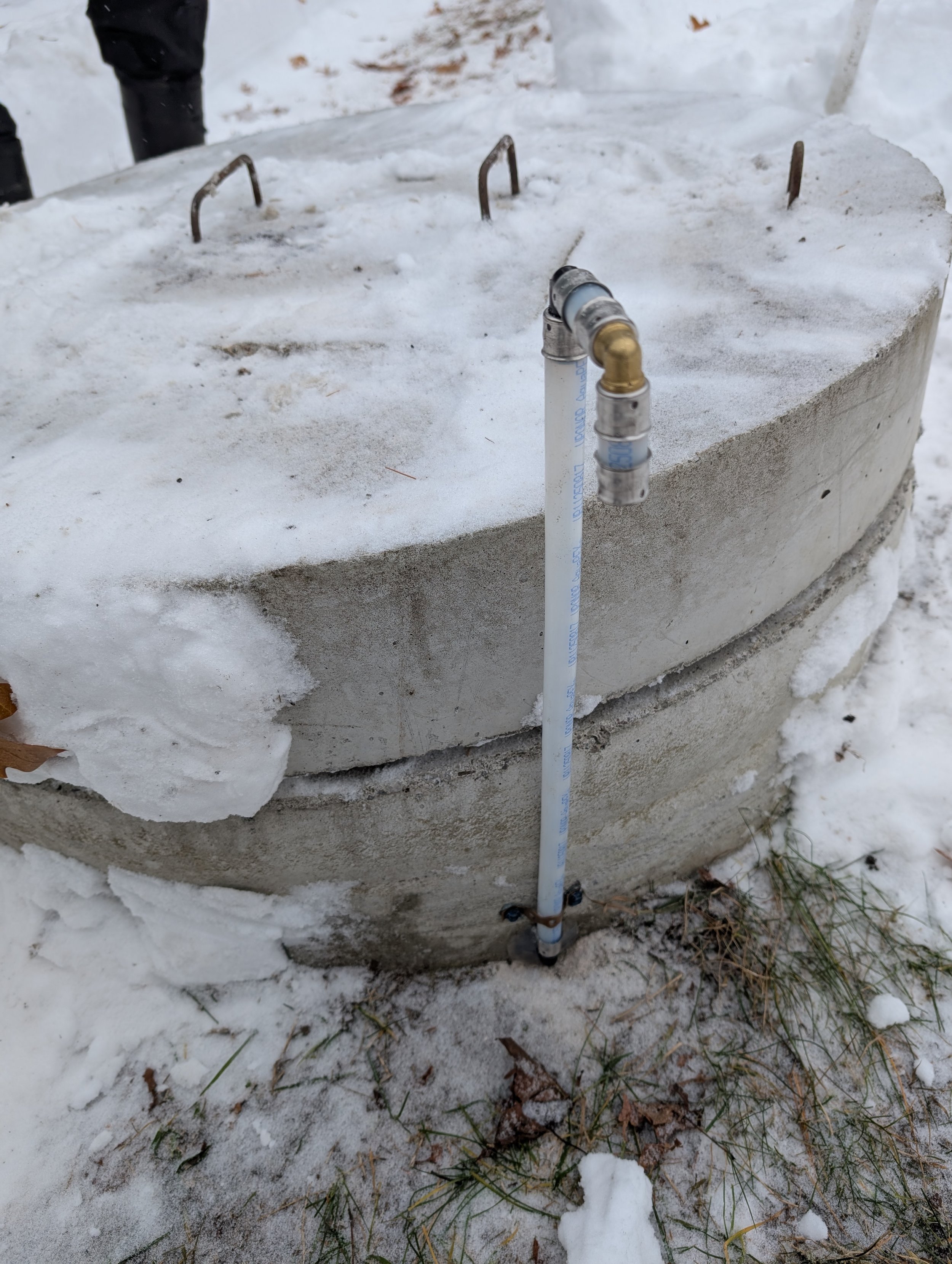 A concrete manhole cover with a metal pipe and a water supply hose attached, surrounded by snow and some grass and leaves on the ground.