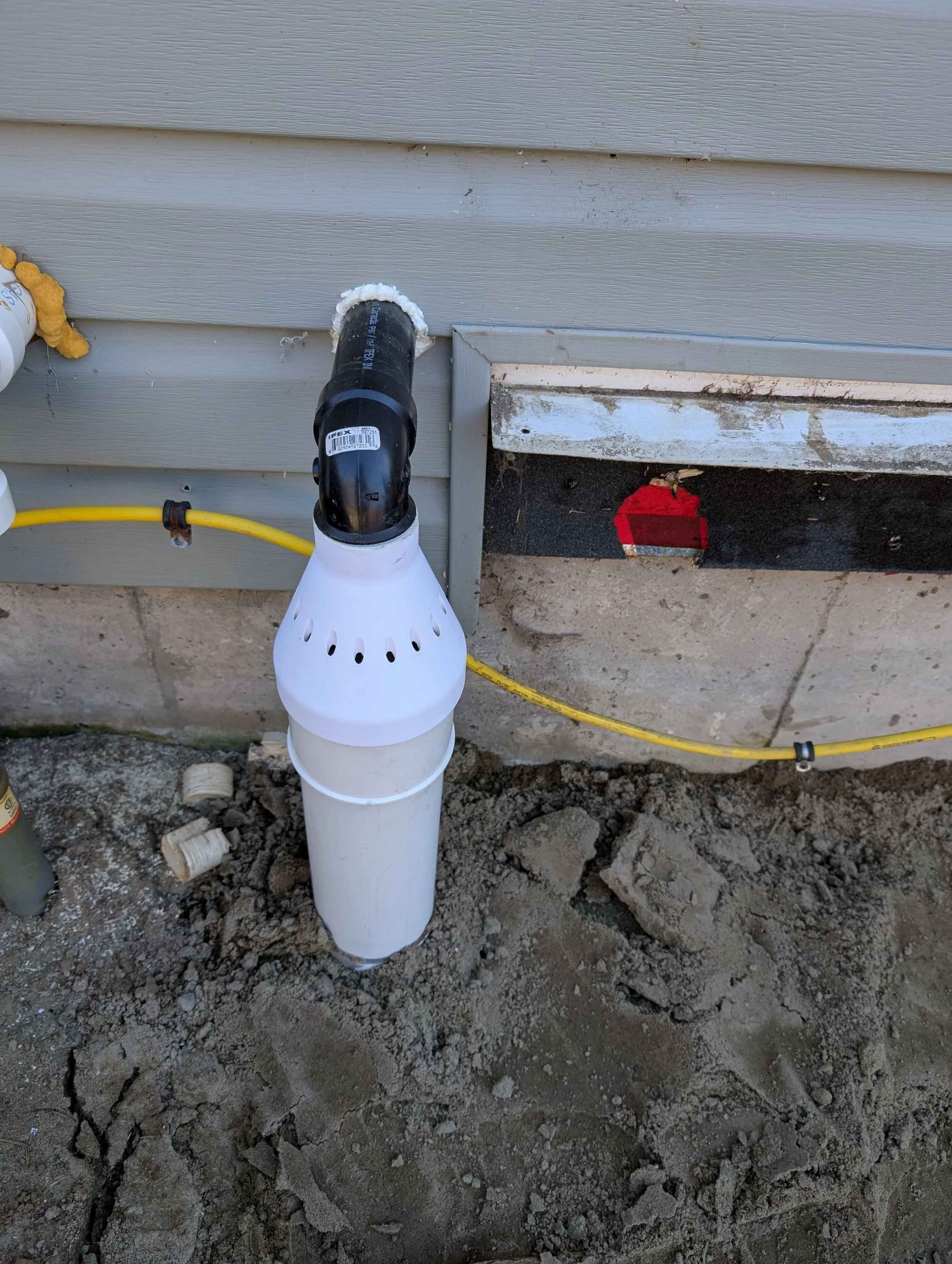 Close-up of a plumbing vent pipe on an exterior wall, with some insulation foam around the pipe, on a construction site with exposed dirt and some debris.