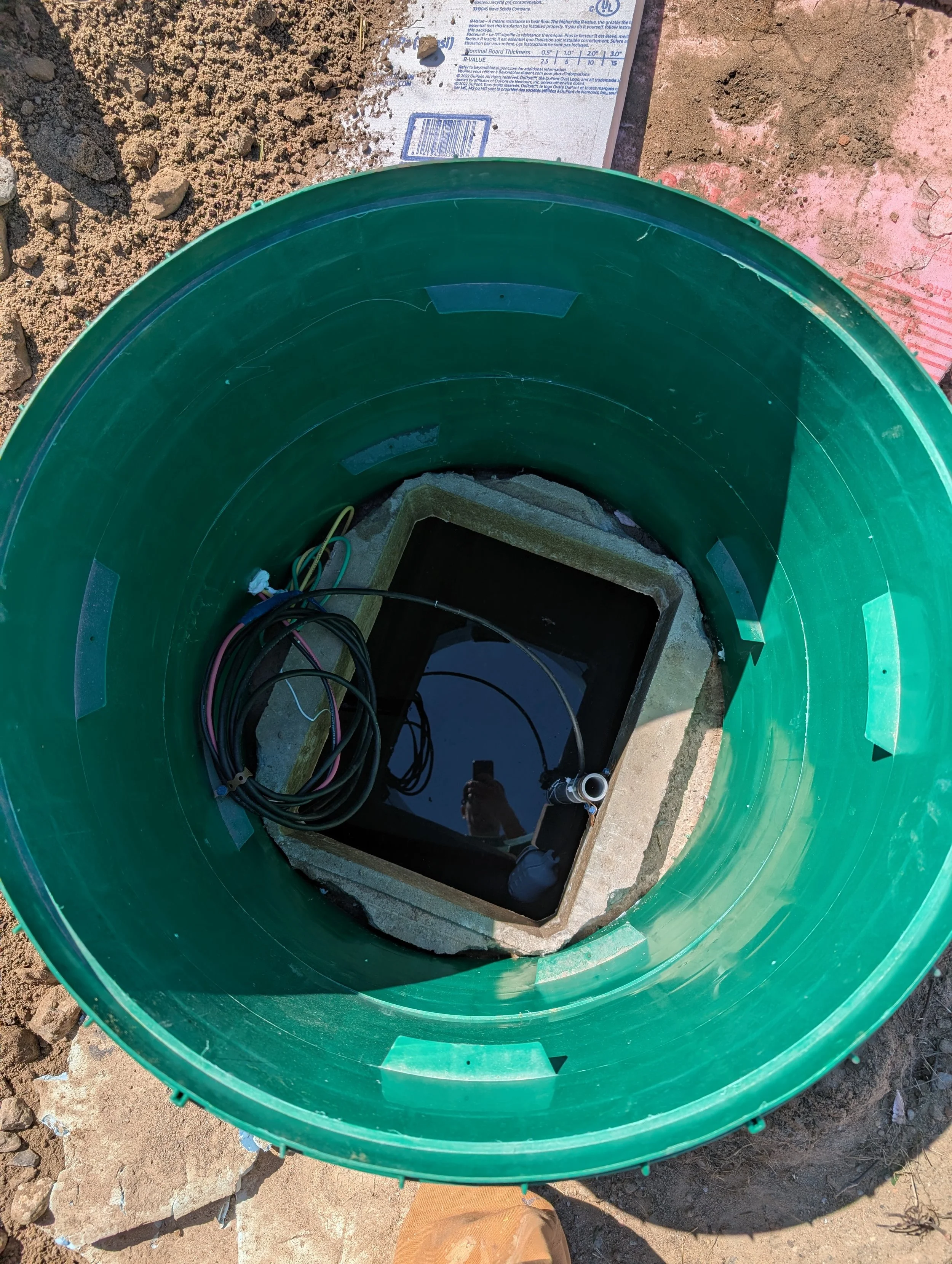 An open green cylindrical inspection chamber or manhole on a dirt ground, showing electrical cables and plumbing inside.
