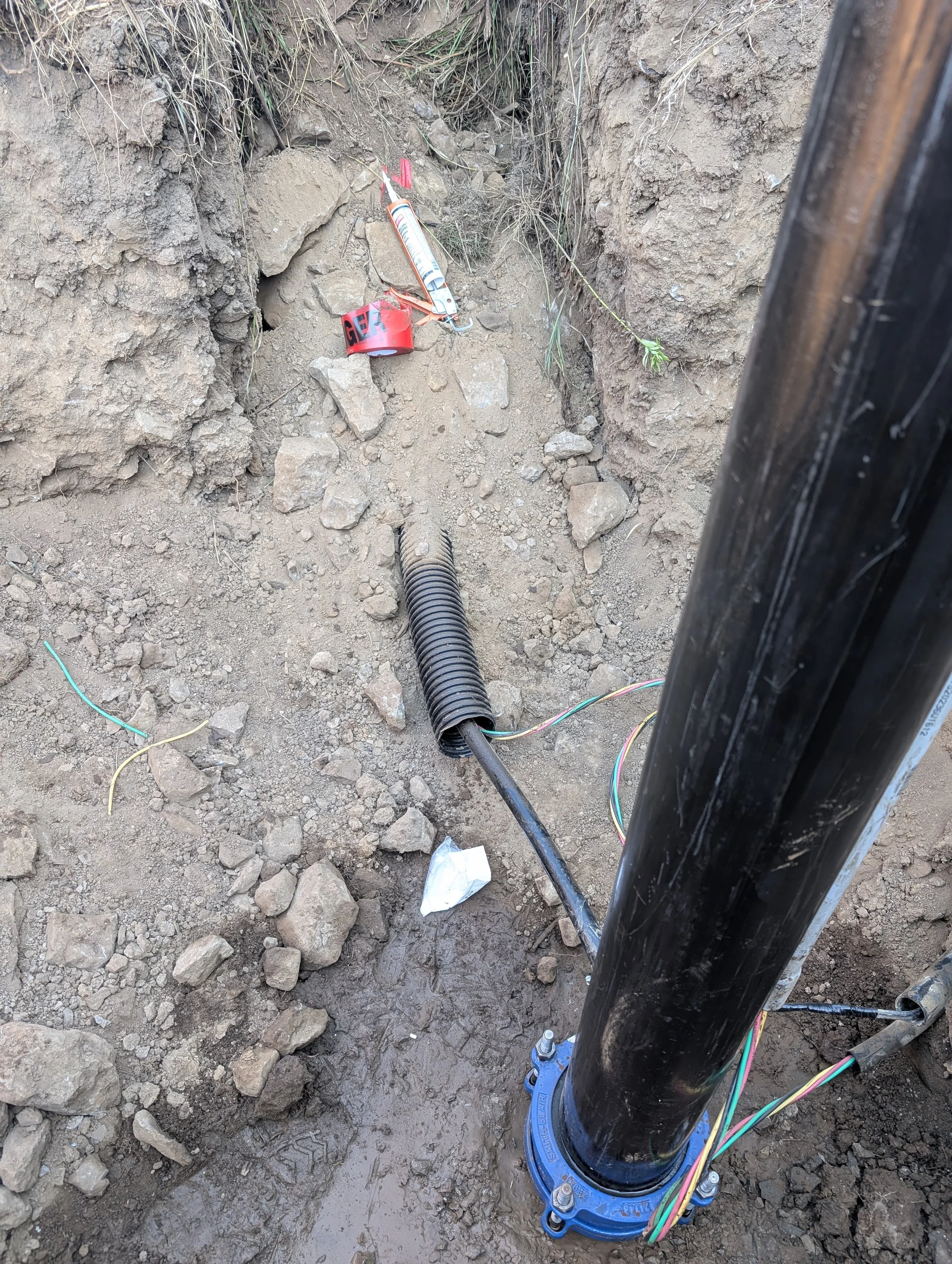 A construction site showing a large black pipe connected to a flexible corrugated pipe, with wires and equipment attached to the area, surrounded by dirt and rocks.