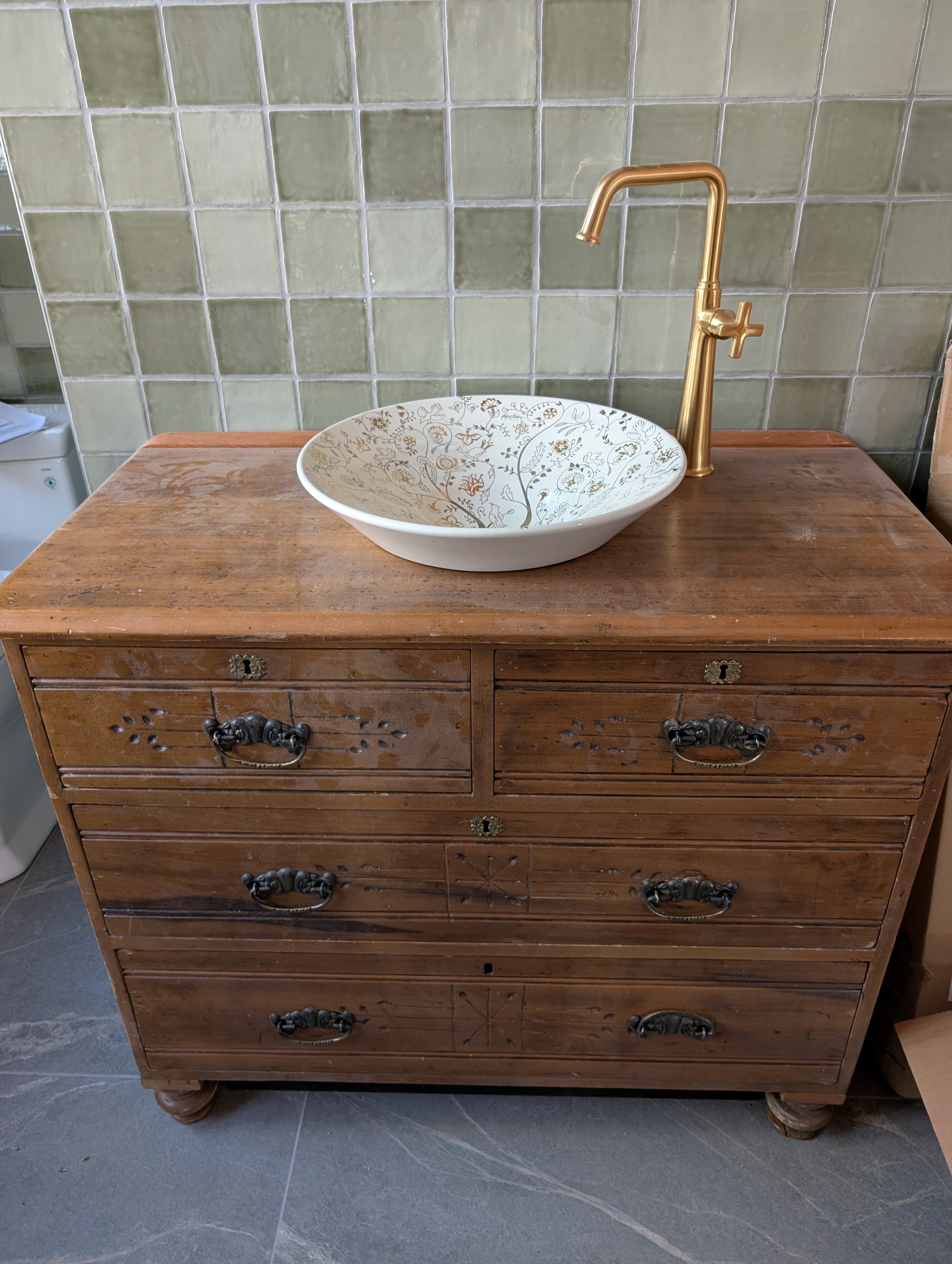 A wooden dresser with multiple drawers and ornate handles, topped with a decorative ceramic bowl containing a gold faucet. The background features a wall with green tiles.