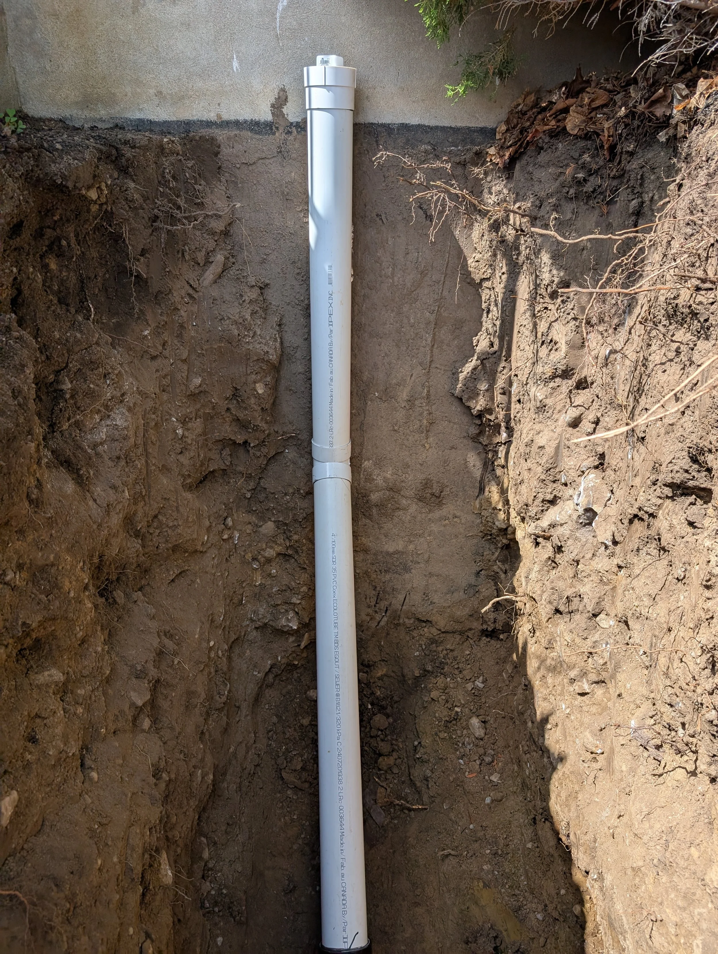 A white PVC pipe installed in a trench with exposed soil on both sides and a concrete wall in the background.