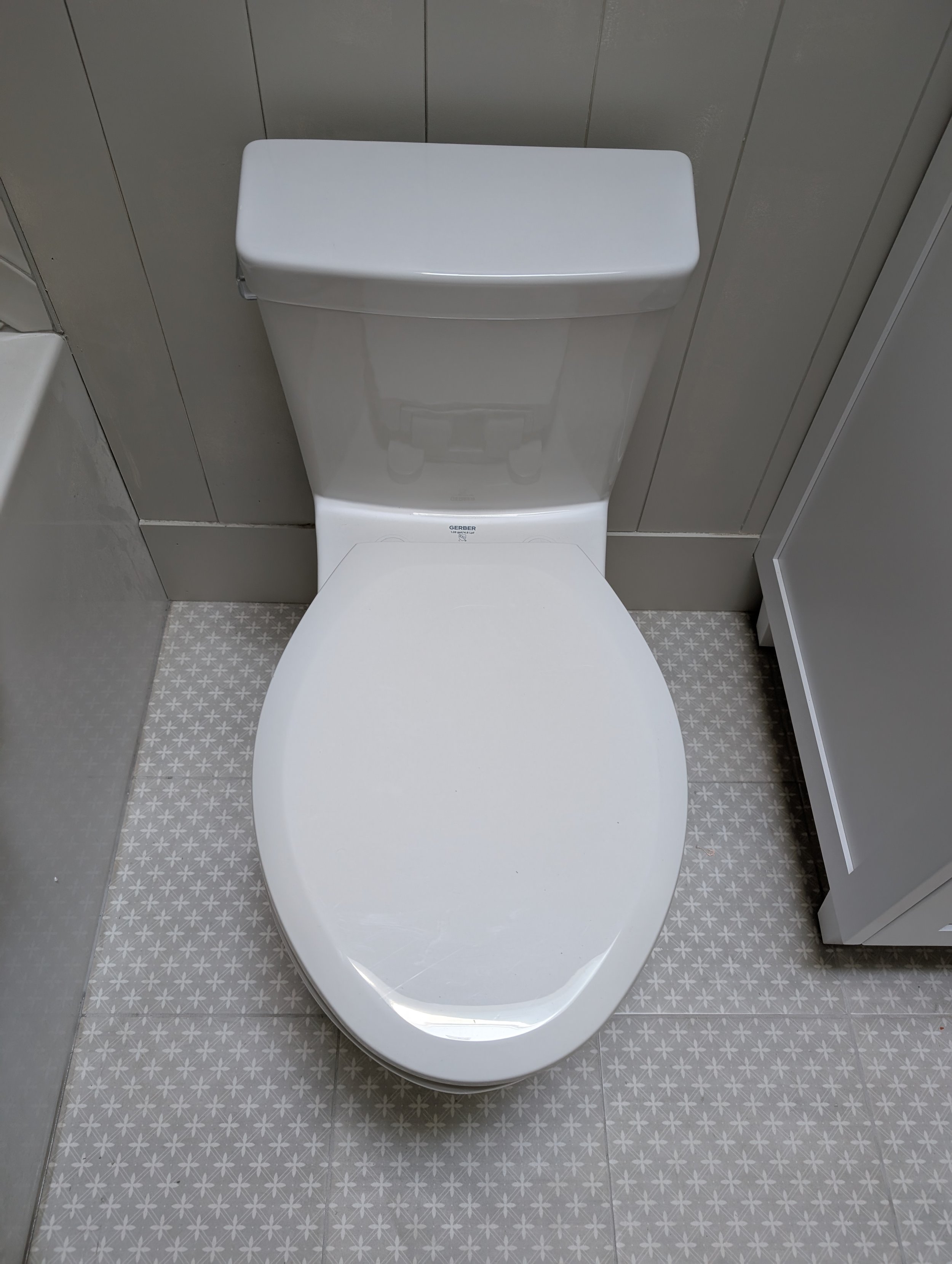 A white toilet with a closed lid in a bathroom, with gray wall panels and patterned tile flooring.