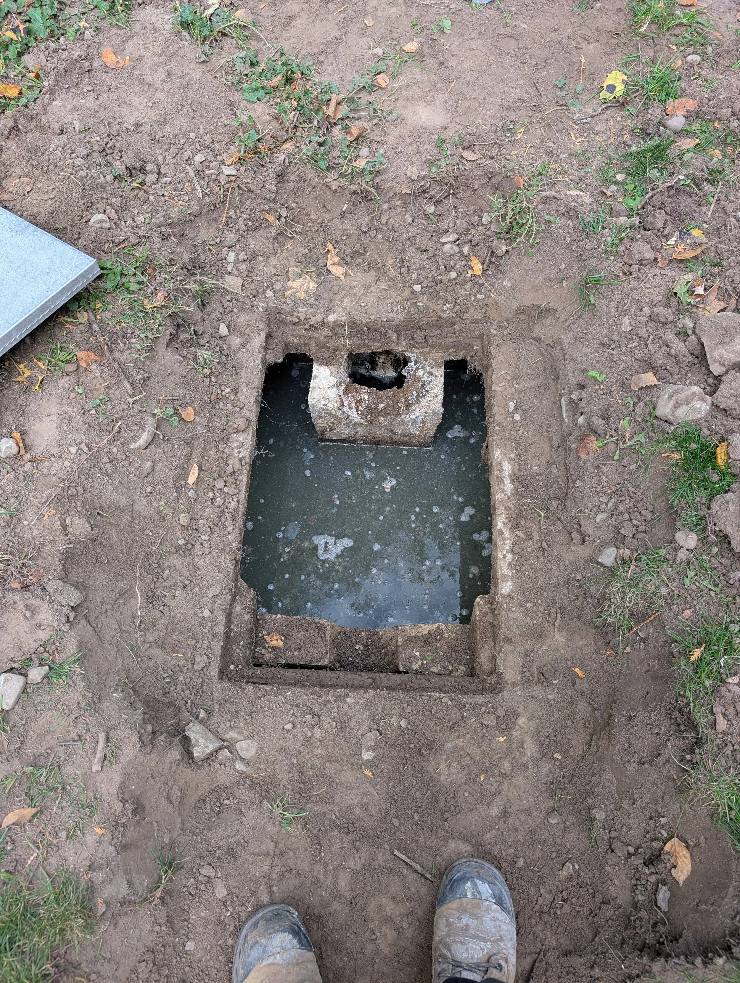 A hole in the ground filled with water, surrounded by dirt and some grass. The photo includes a person's muddy boots at the bottom.
