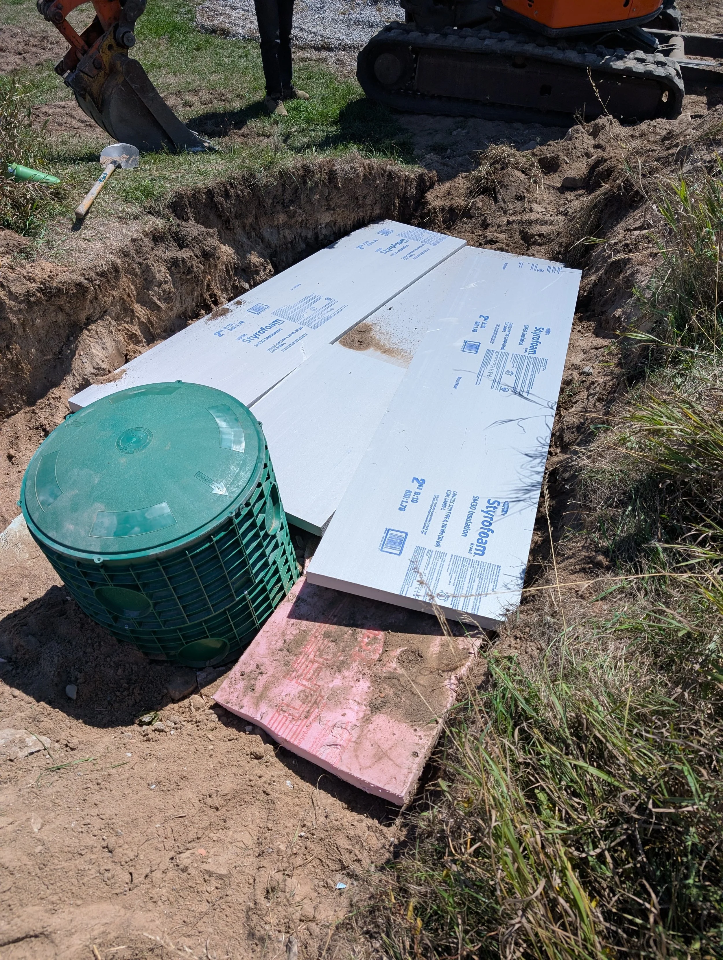 A construction site dugout with Styrofoam insulation panels placed at the bottom of the trench. An excavator is nearby, and construction tools are on the ground. The area is outdoors with dirt and sparse grass.