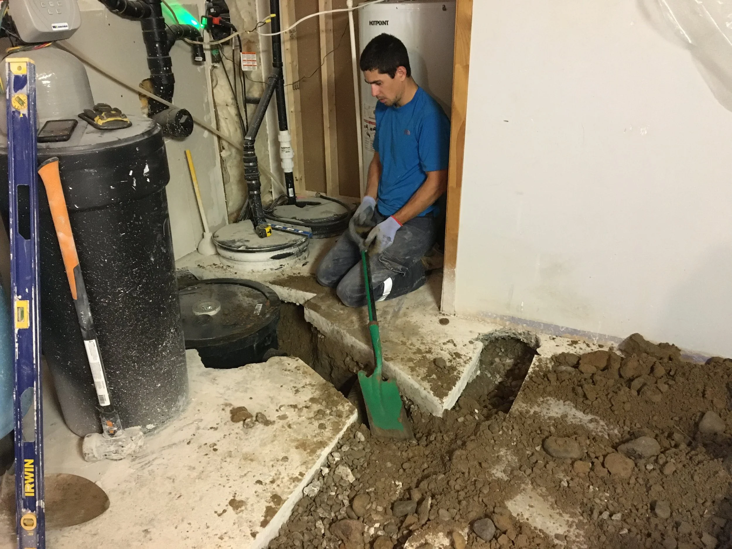 A plumber is working on a home improvement project in a basement. He is kneeling beside a deep hole in the concrete floor, with a green shovel nearby. The area appears to be under renovation, with exposed plumbing, insulation, and unfinished walls.