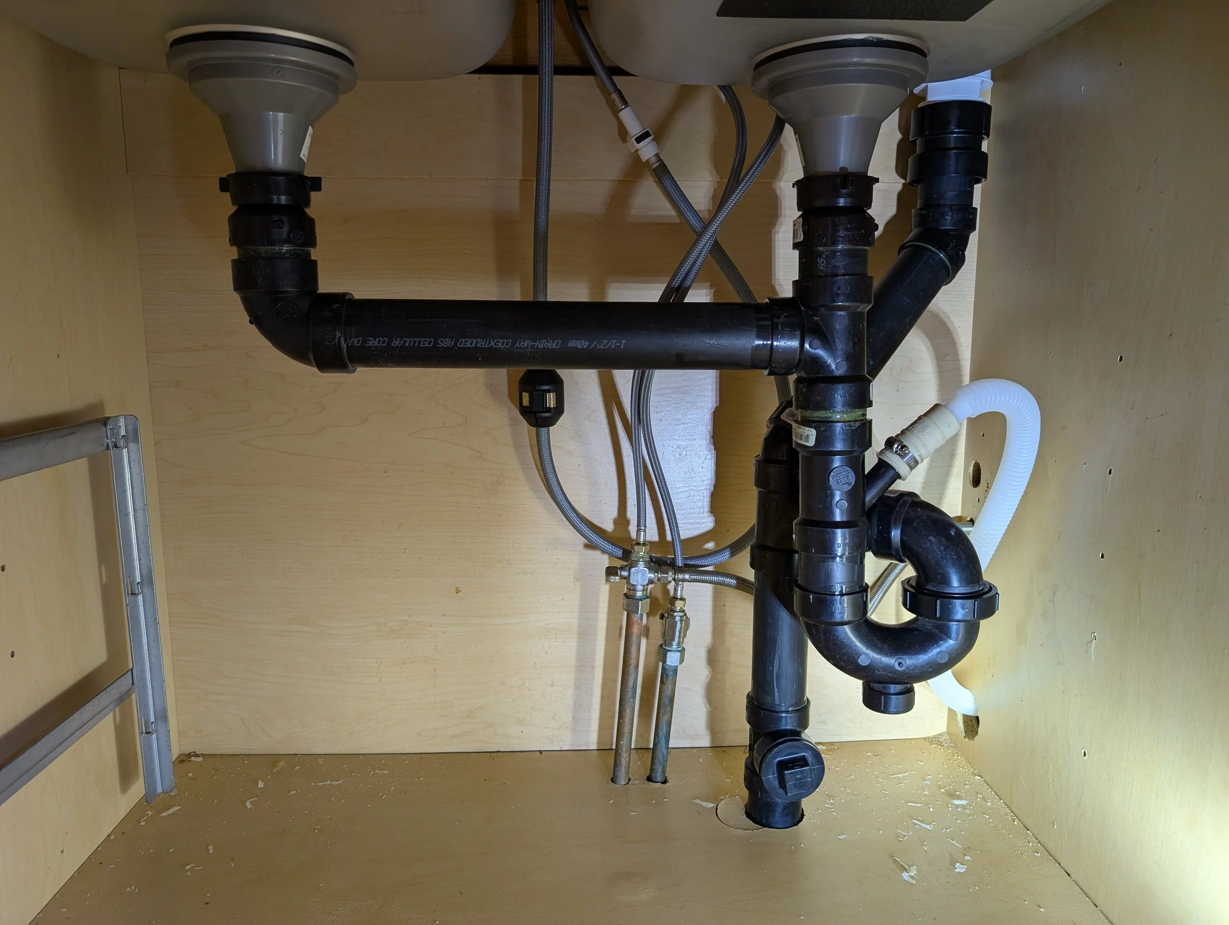 Under-sink plumbing with black pipes, metal hoses, and PVC fittings inside a wooden cabinet with a small ladder on the left.