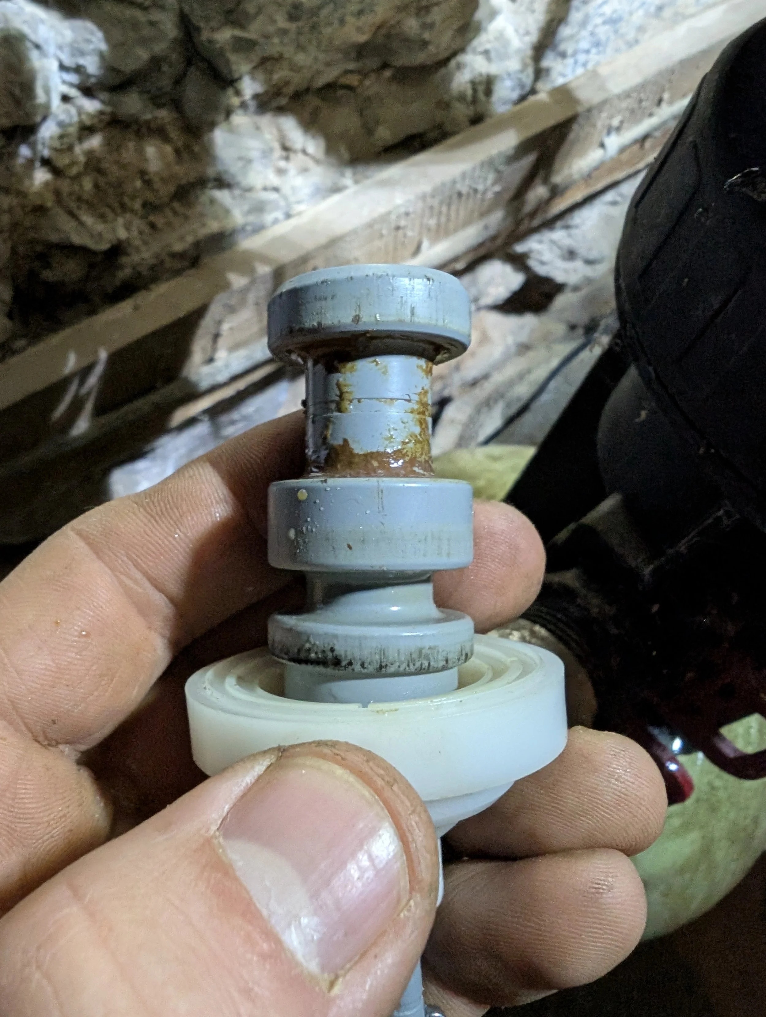 Close-up of a person's hand holding a small mechanical part, possibly a valve or a pipe component, in front of a brick and stone wall background.