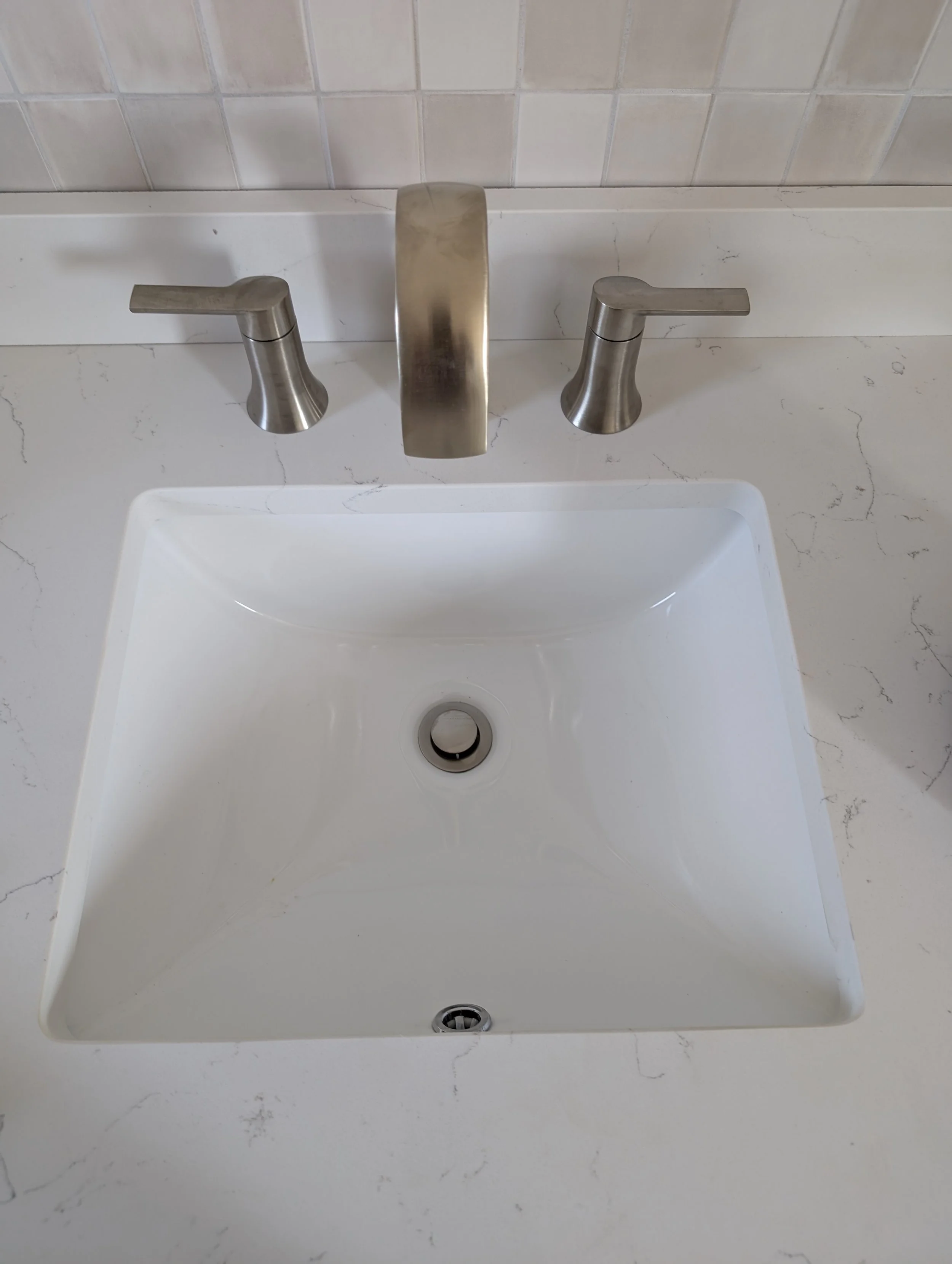A white rectangular bathroom sink with a stainless steel faucet and handles. The background features a tiled wall with beige and light brown tiles. The sink drain is visible at the bottom center.