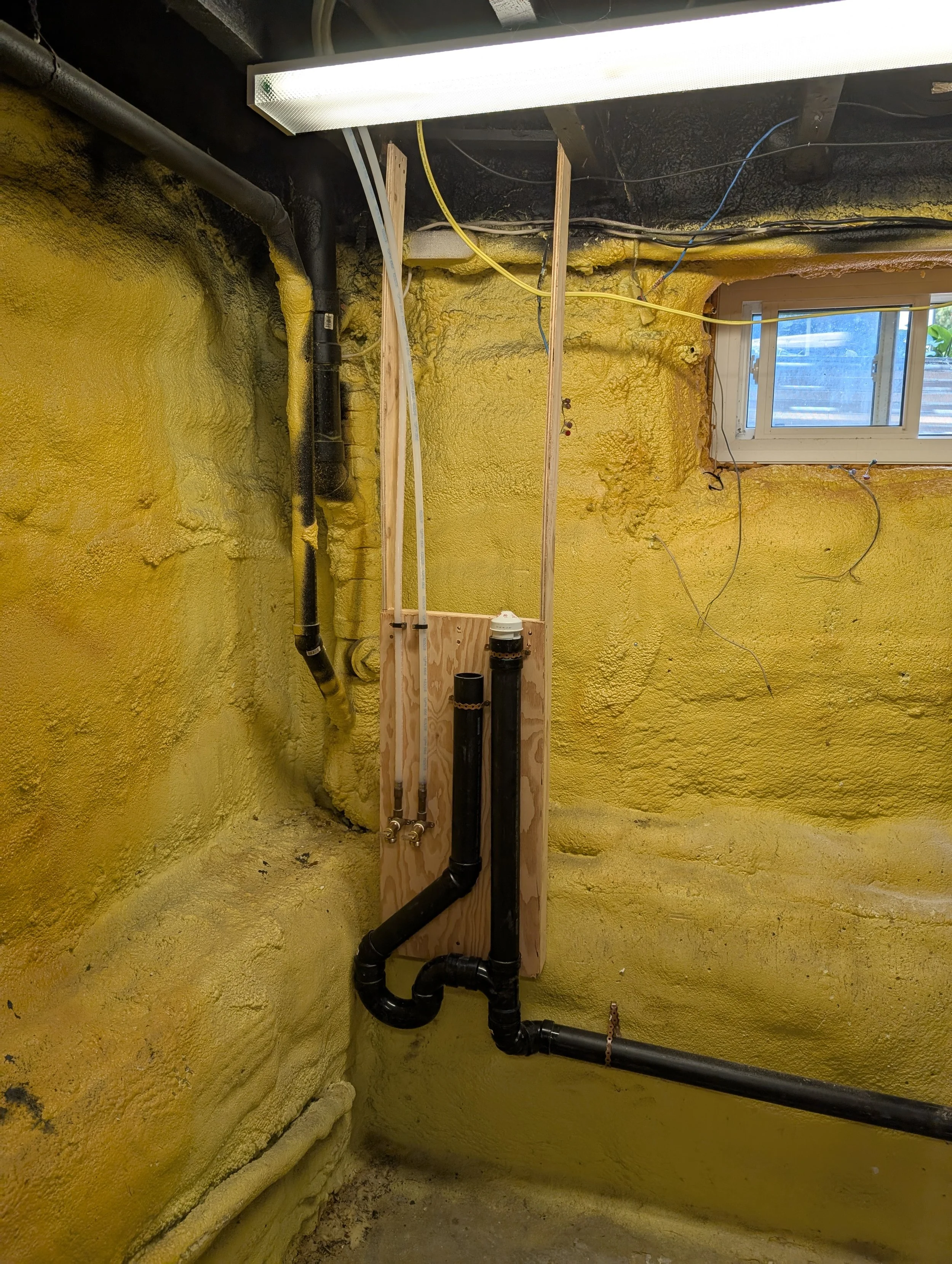 Interior of a basement with yellow spray foam insulation on the walls, black and black pipes running vertically and horizontally, and a small window on the right wall. There are exposed wires, a wooden frame, and a fluorescent light fixture on the ce