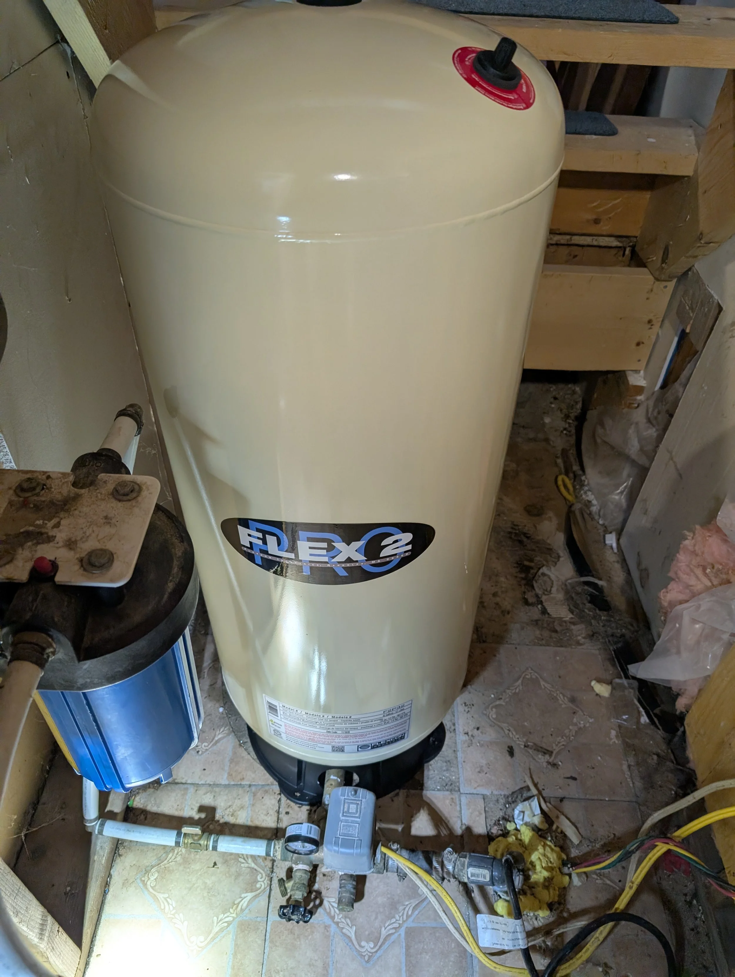 A beige Rheem water heater installed in a basement or utility room, with various plumbing and electrical connections, surrounded by exposed wooden framing and construction materials.