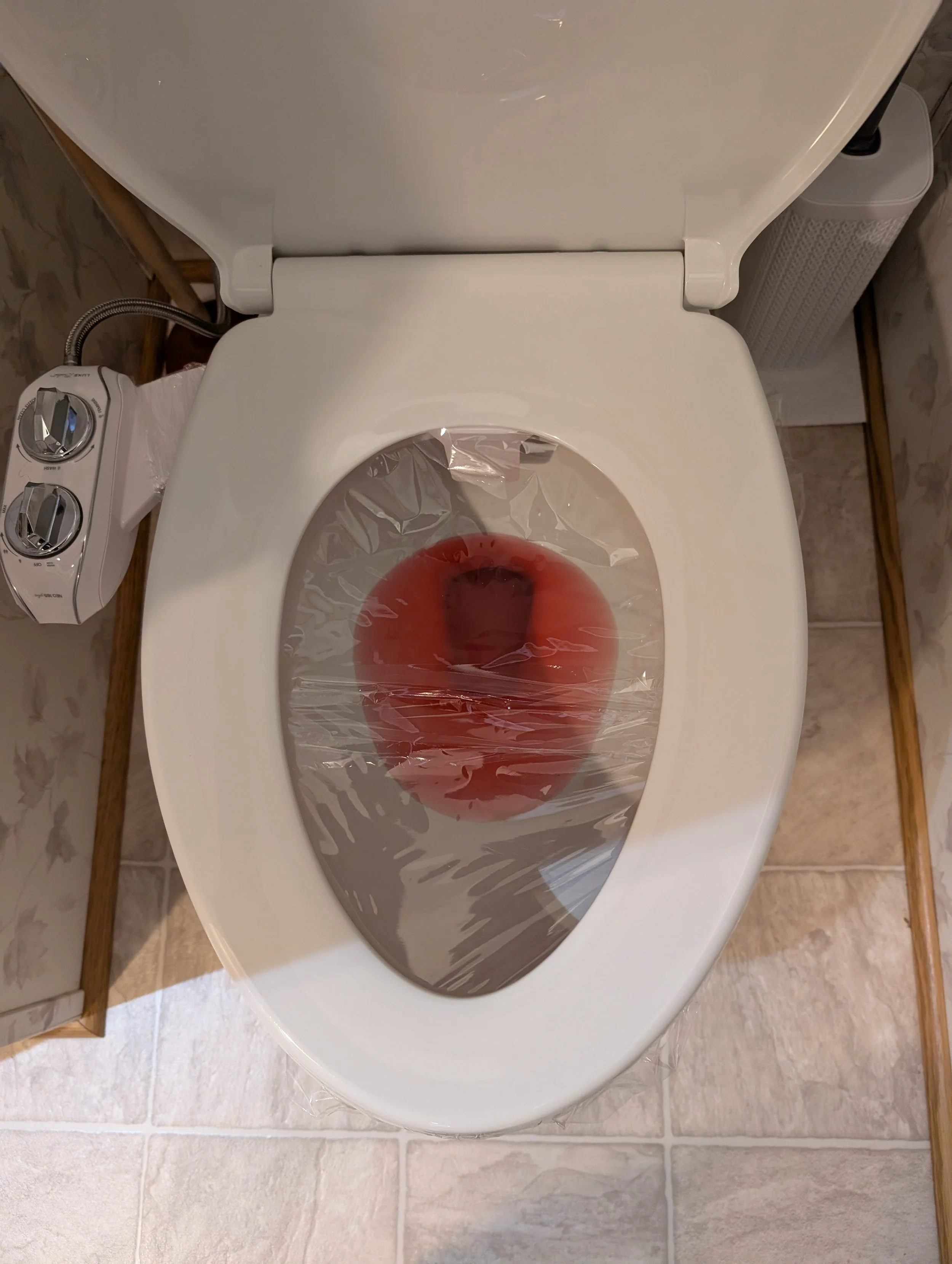 A white toilet with the lid open, placed on a tiled bathroom floor. The toilet seat is covered with a plastic liner containing red liquid, possibly for sanitation or protection.