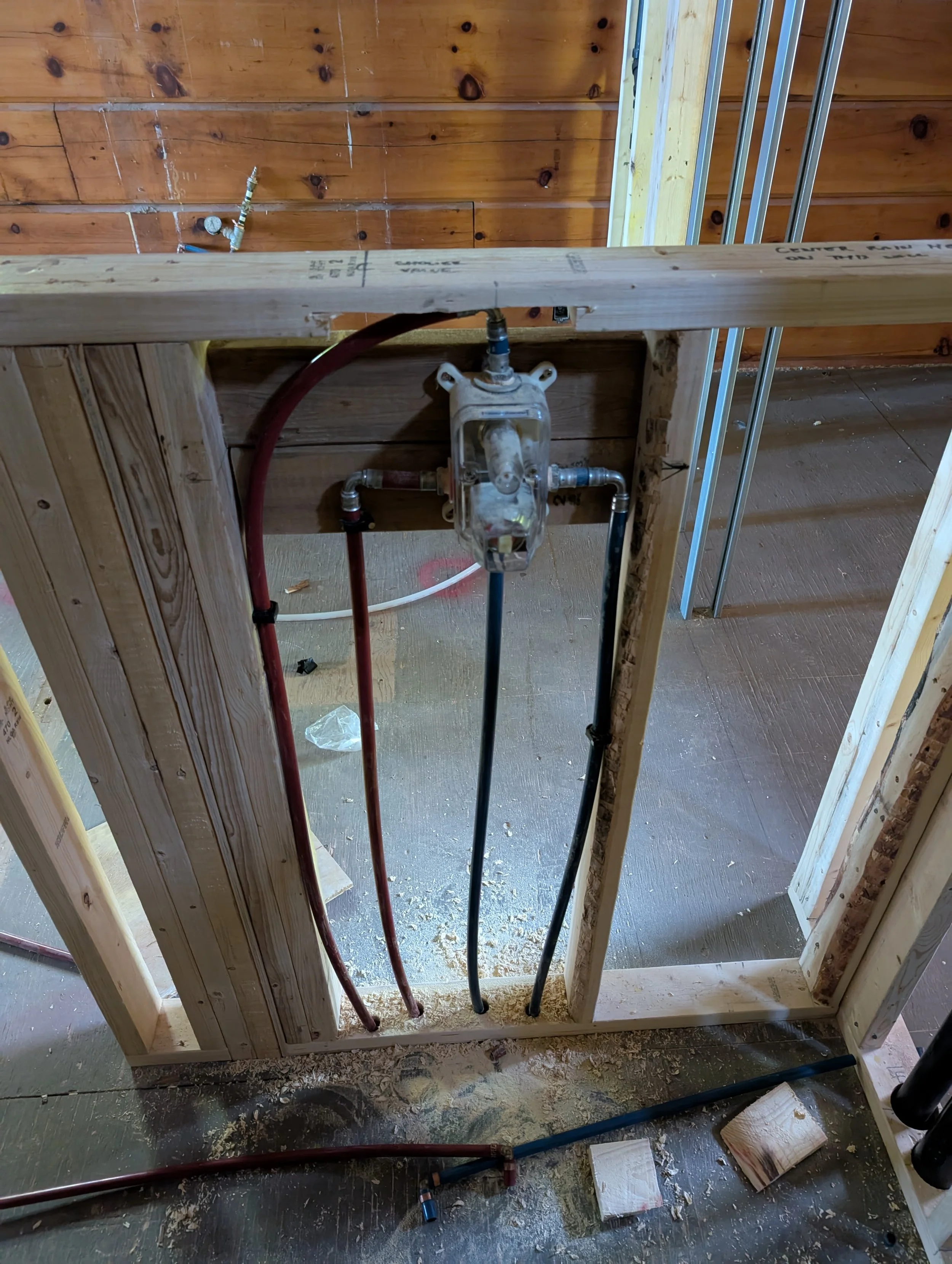 Interior of a building under construction featuring exposed pipes, wooden framing, and an unfinished wall. Construction materials and debris are on the floor.