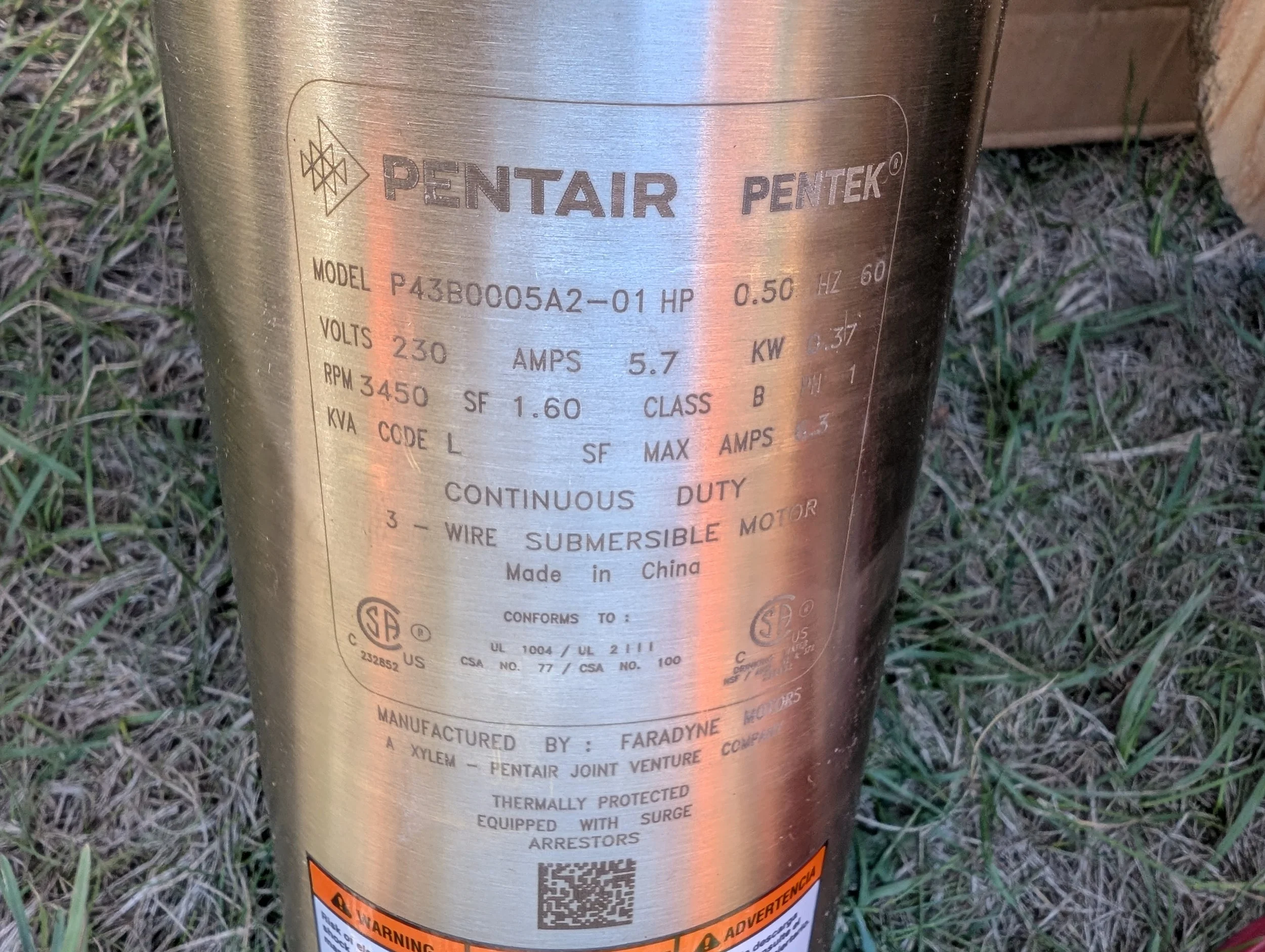 A metallic cylinder with a label containing technical specifications, brand names PENTAIR and PENTEK, and warning signs, placed outdoors on grass.