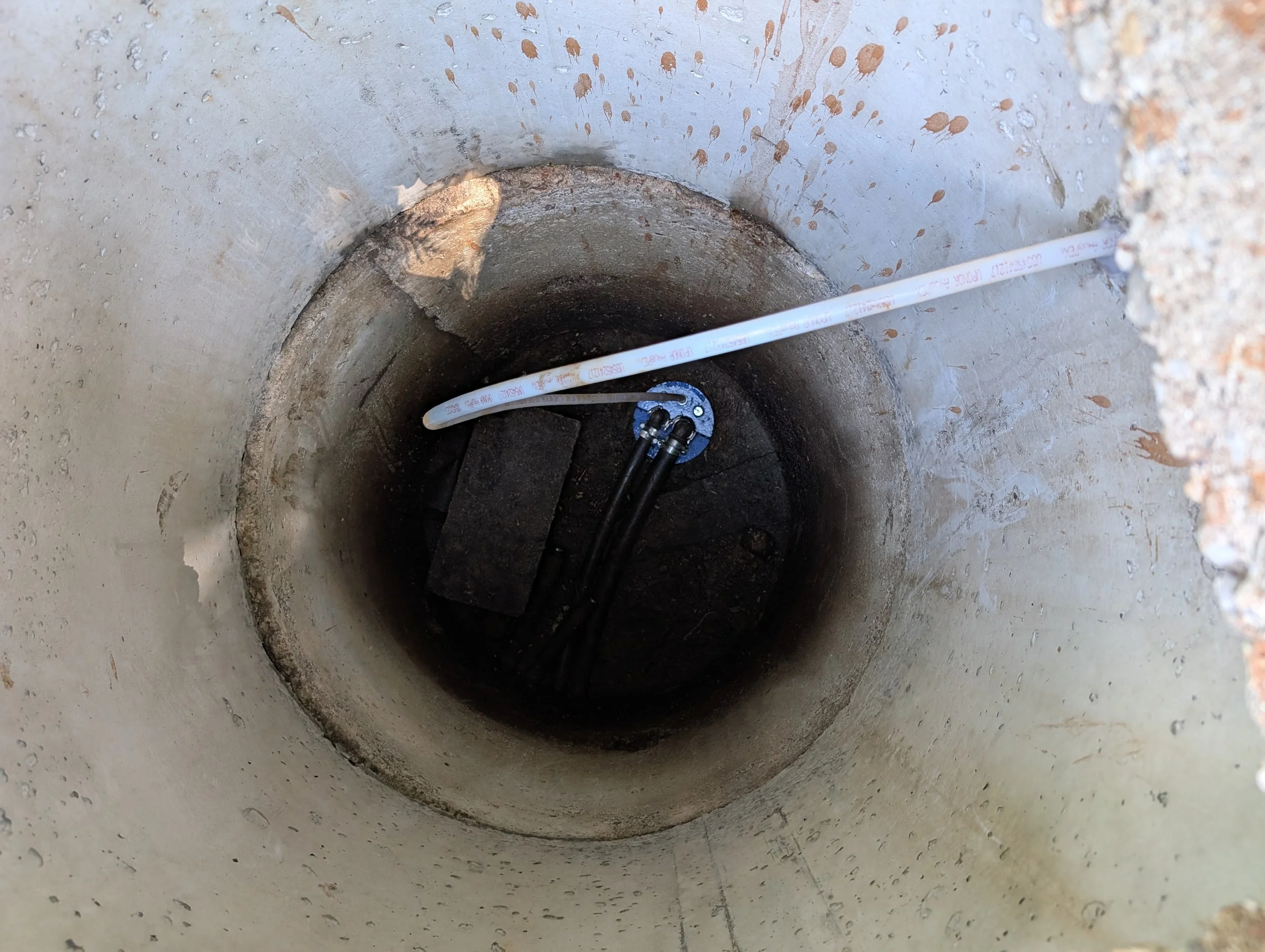 A deep hole in the ground with a white pipe running through it, connected to a blue metallic fixture, with small dirt and debris around the hole.