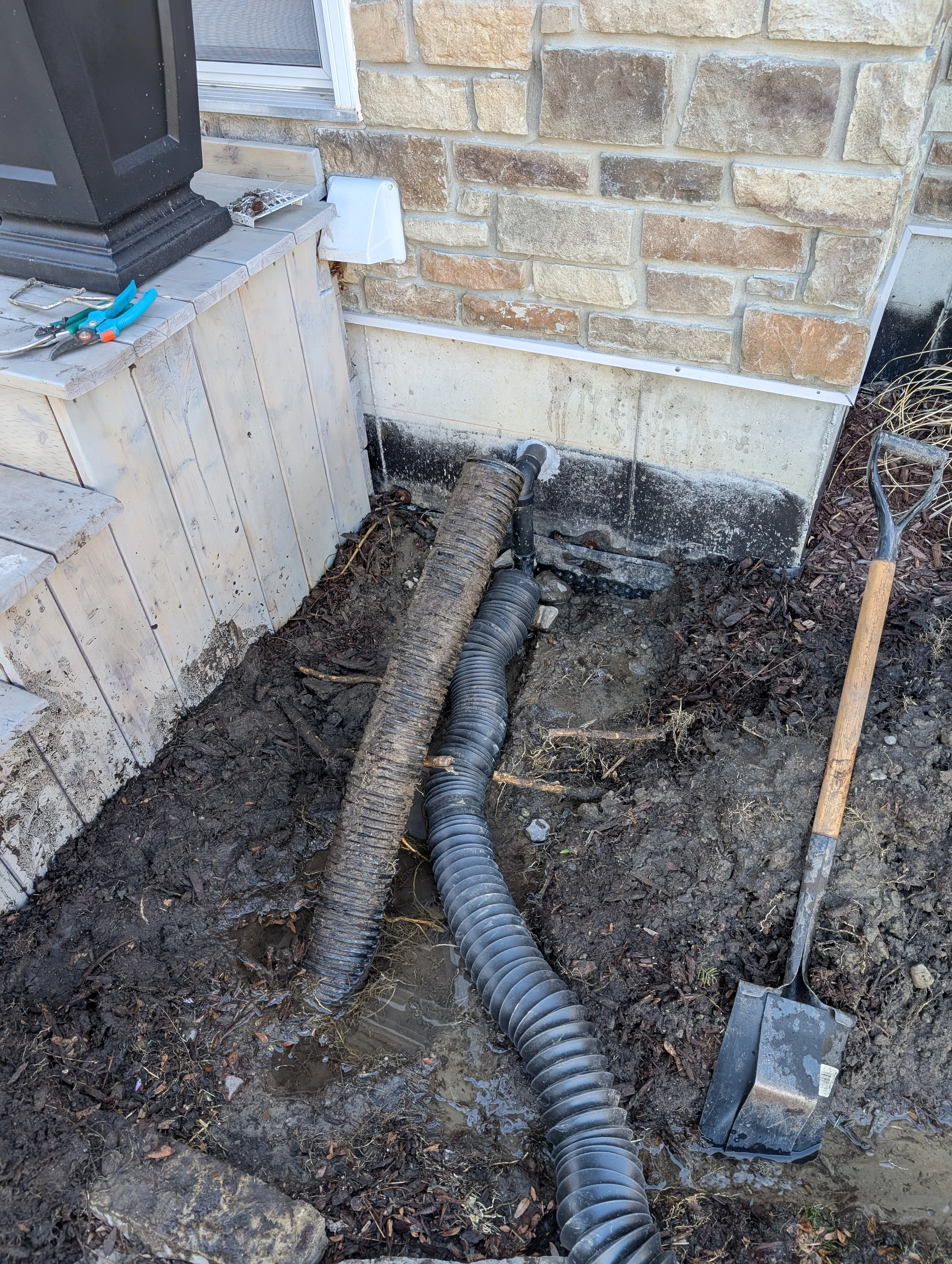 A construction site showing a large black drainage pipe connected to a chimney vent, with tools and a black plastic cover nearby, and a wooden deck with a small overhang on a brick house exterior.