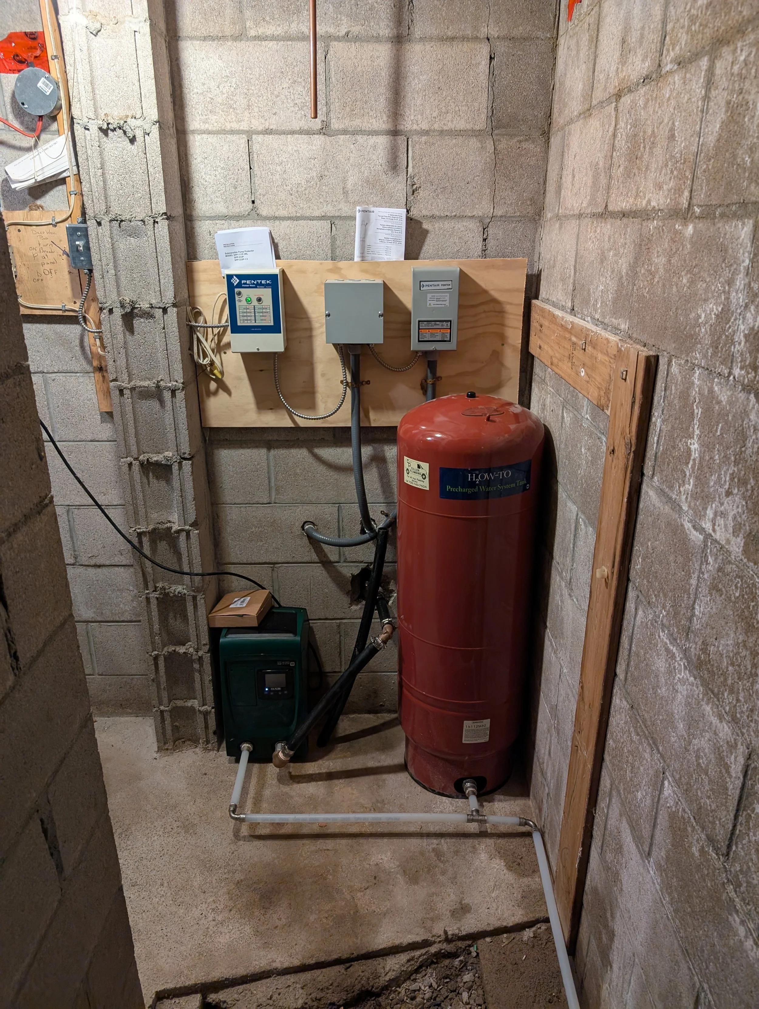 A utility space with various electrical and plumbing components including a water pressure tank, control boxes, pipes, and unfinished cinder block walls.