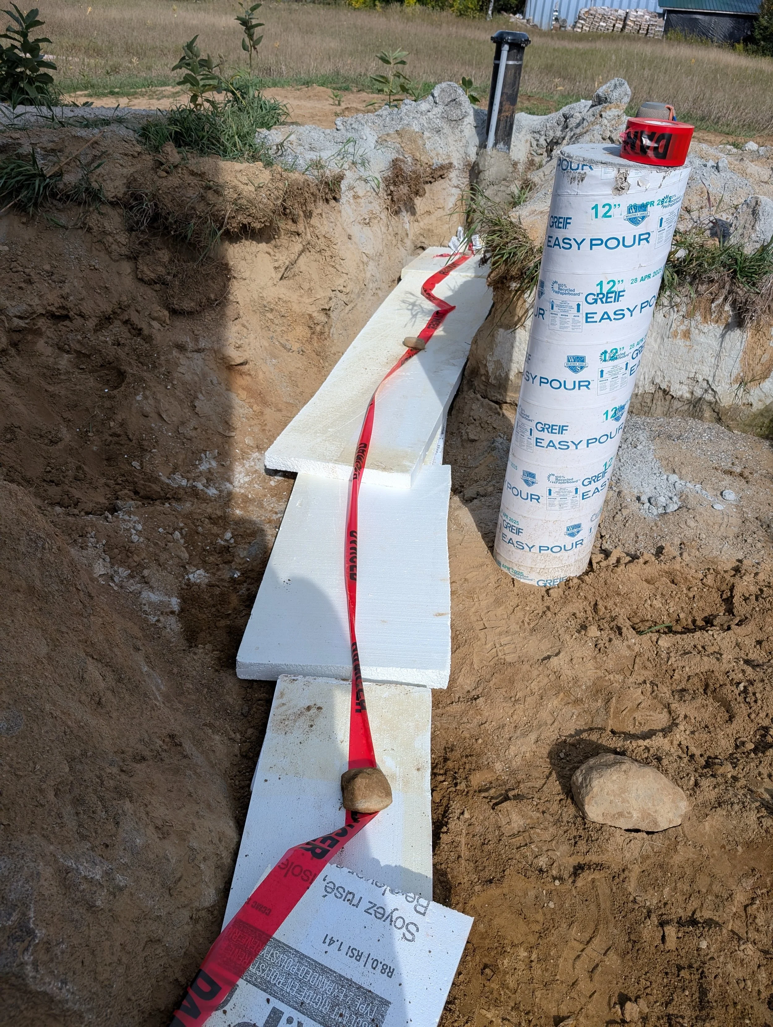 Construction site featuring a large hole in the ground with a white insulating foam board placed inside, a red caution tape, a large roll of weather-resistant paperboard, and a small black pipe protruding from the ground