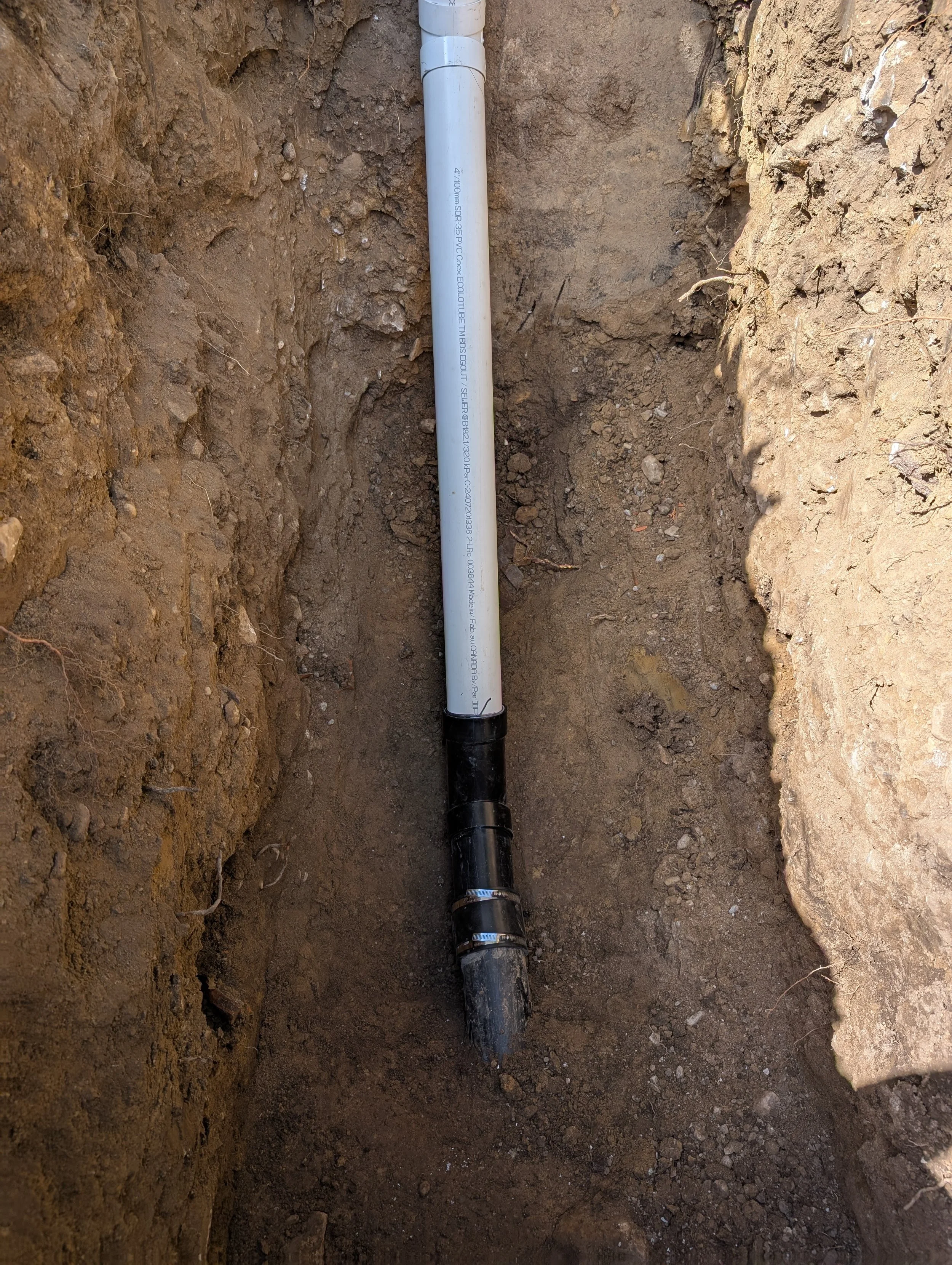A PVC pipe laid in a freshly dug trench in the dirt, with pipe fittings visible at the top and bottom.