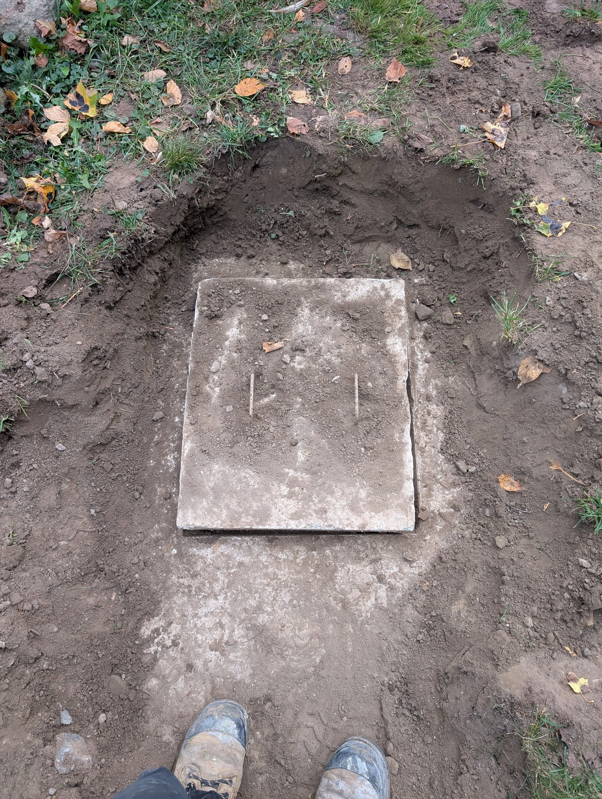 Person wearing muddy shoes standing on dirt and grass with a rectangular stone at their feet and a hole in the ground.