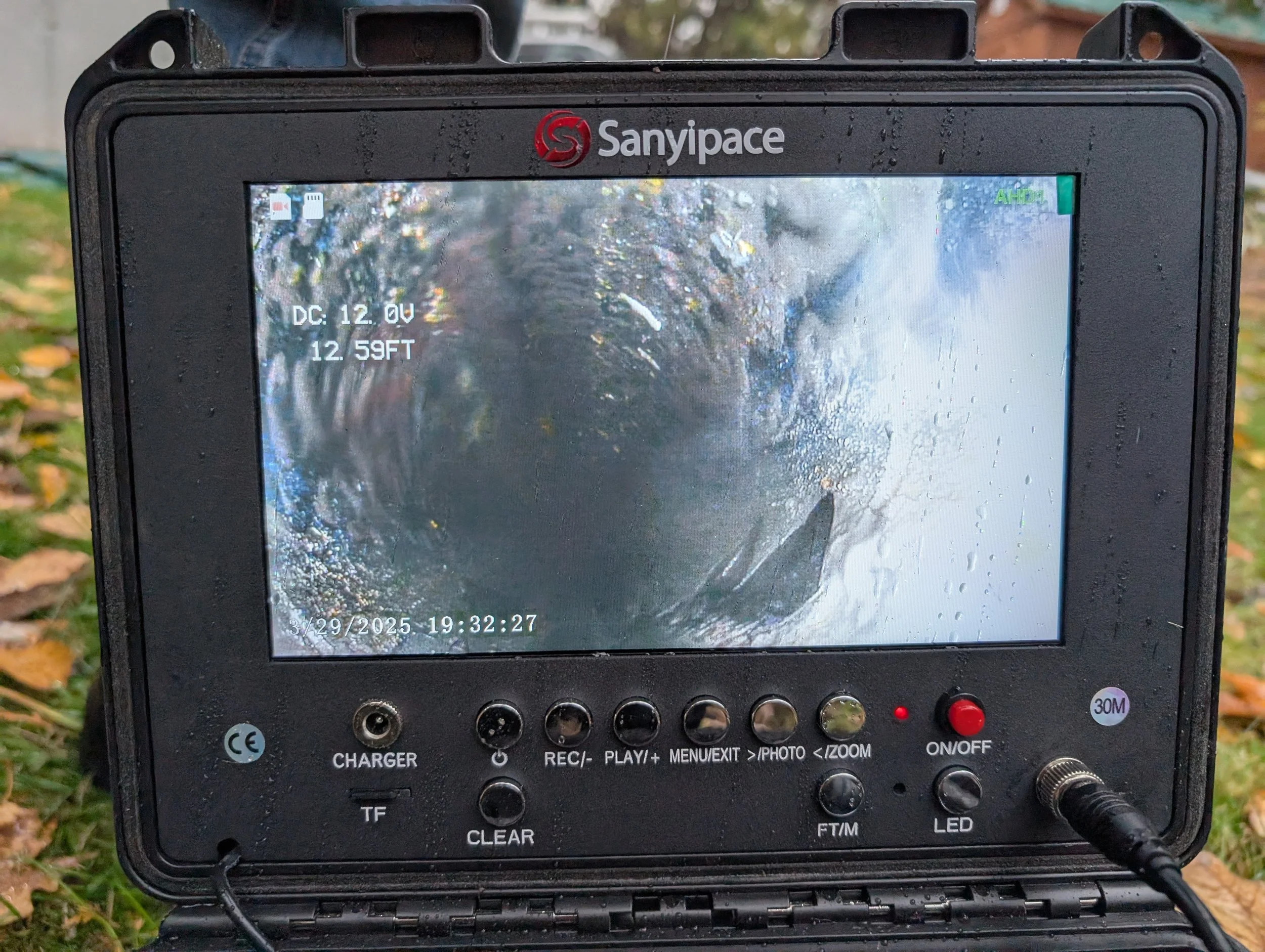 A black electronic device with a screen showing a water splash scene, located outdoors on grass with leaves. The device has various labeled buttons and ports, with text indicating voltage, date, time, and functions.