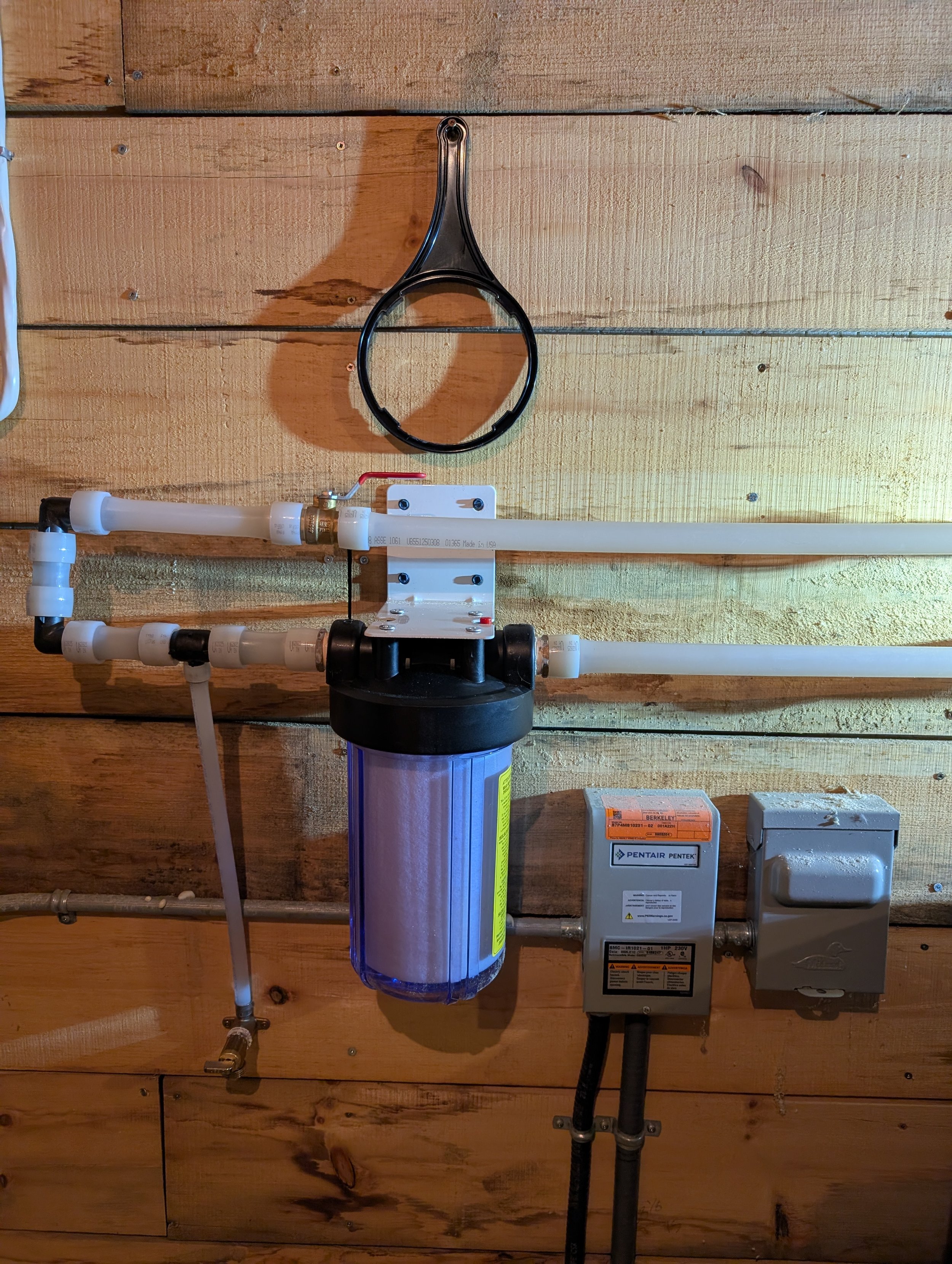 Water filtration system mounted on a wooden wall, with PVC pipes, a large water filter, and electrical components.
