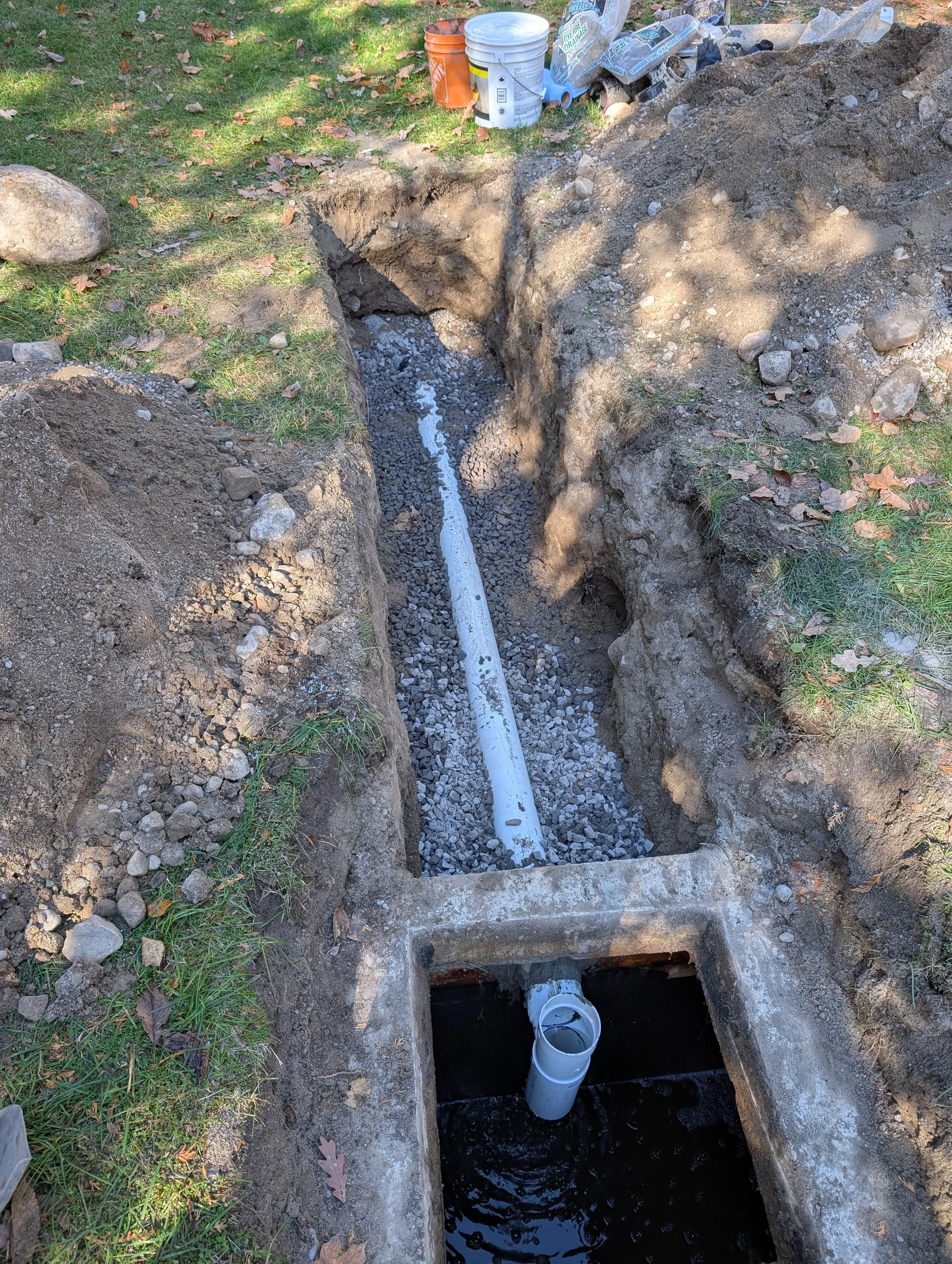A construction site with an open trench for underground piping, with a white pipe visible inside the trench and construction materials and tools nearby.
