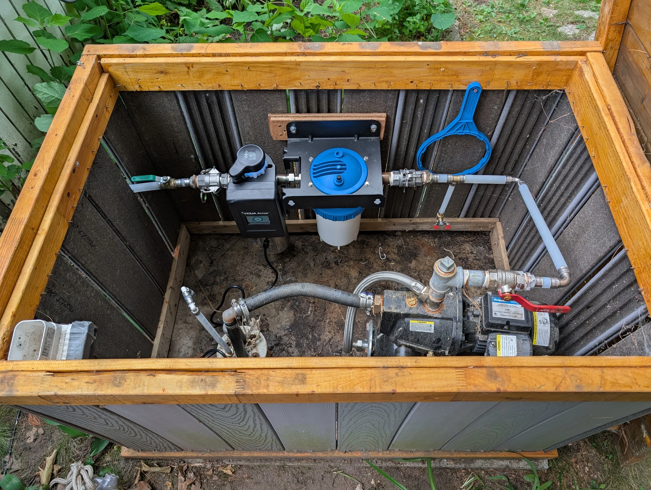Open framework with water filtration system and plumbing pipes, installed in a wooden structure with greenery background.