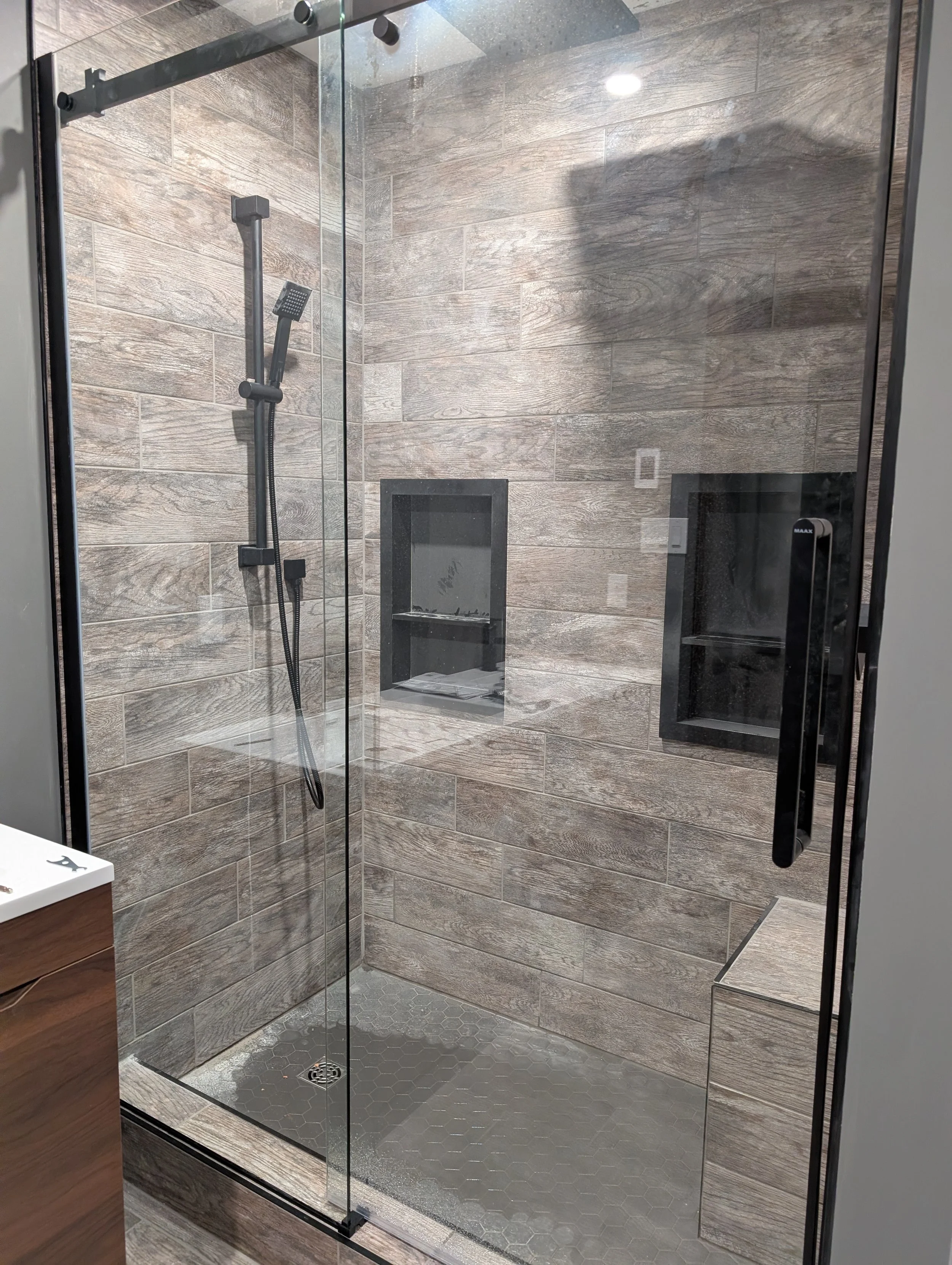 A modern shower enclosure with wooden textured wall tiles, a black shower panel with adjustable hand shower, and a built-in shelf for toiletries. The shower has a glass door and floor with hexagonal tiles.