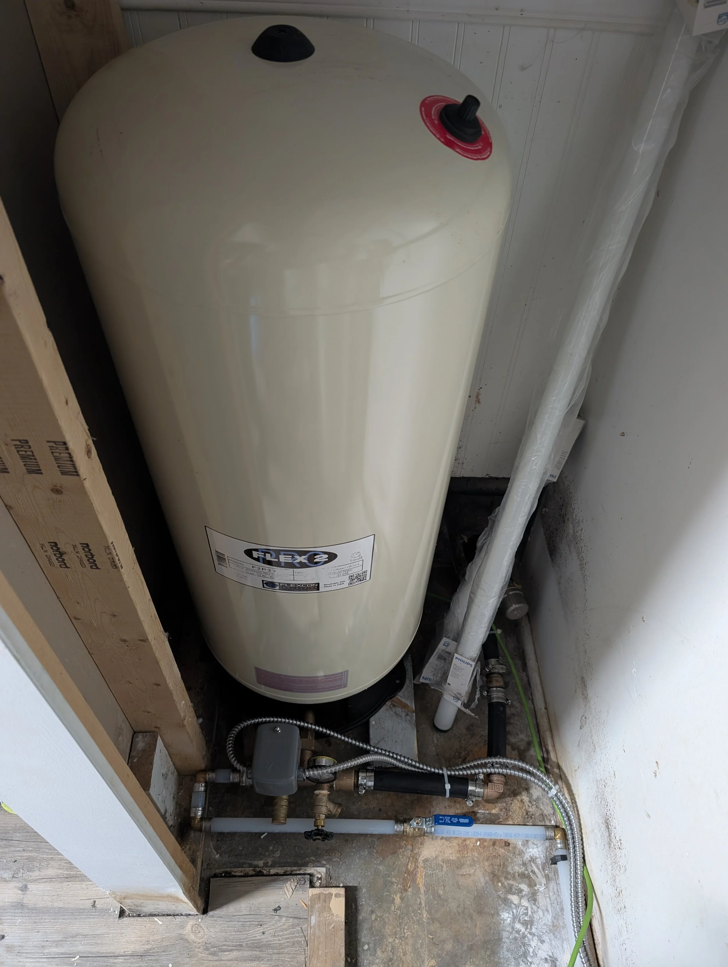 A white, cylindrical water heater installed in an unfinished space with exposed pipes, valves, and electrical connections below.