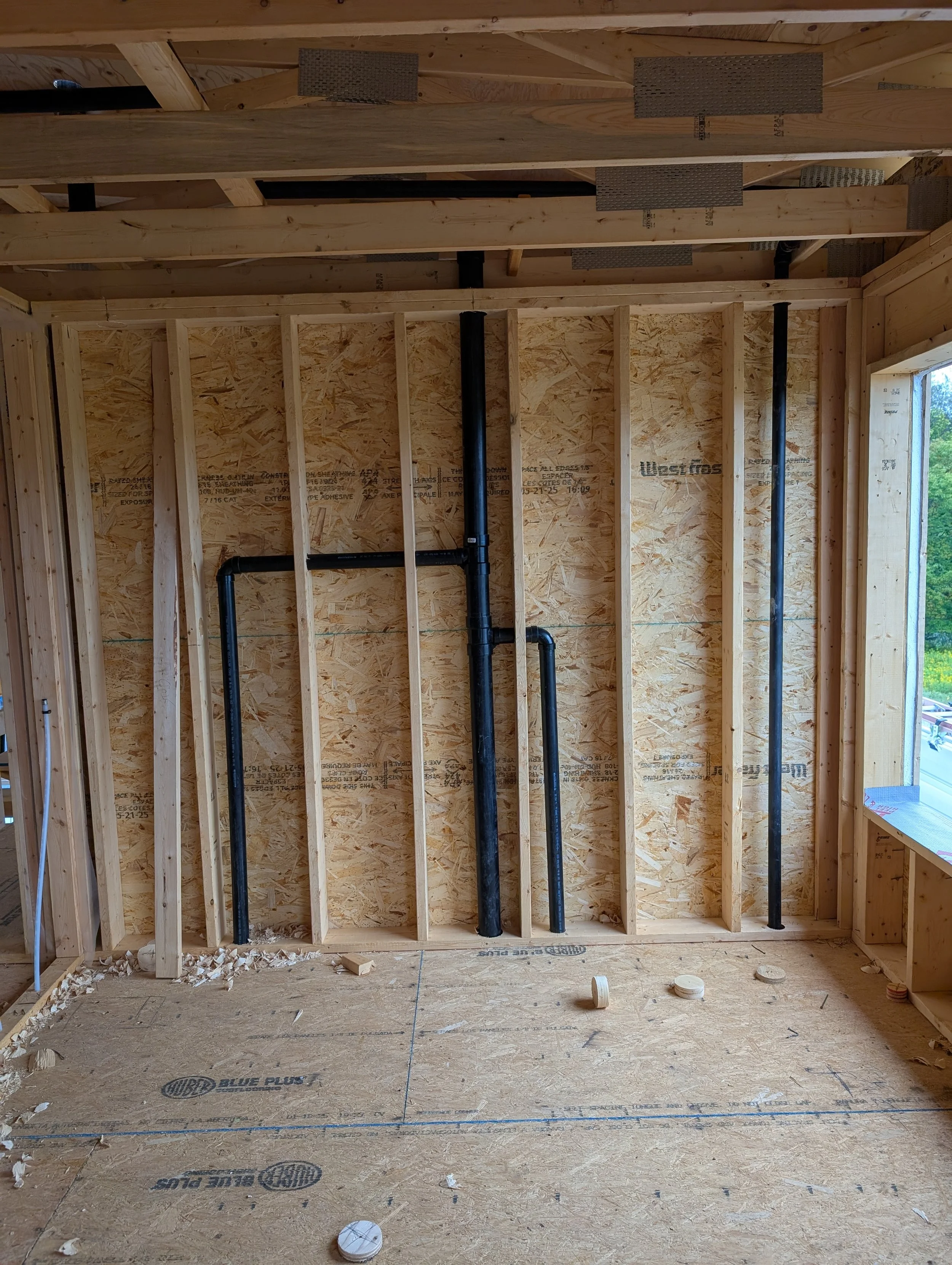 Interior view of a construction site showing wooden framing, black plumbing pipes, and construction debris on the floor.