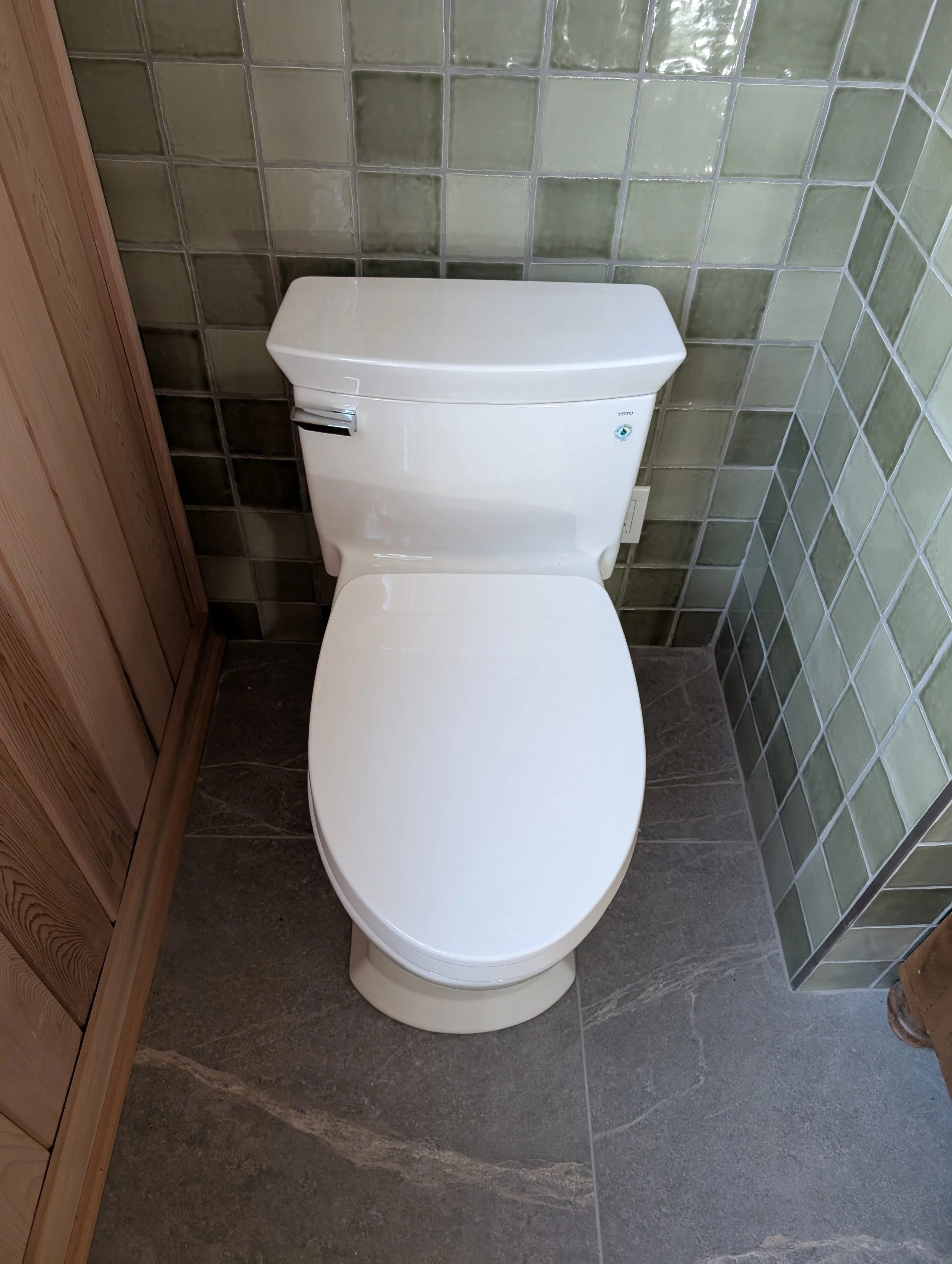 A white toilet in a bathroom with gray tile flooring and green tiled walls, partially covered by a wooden cabinet on the left side.