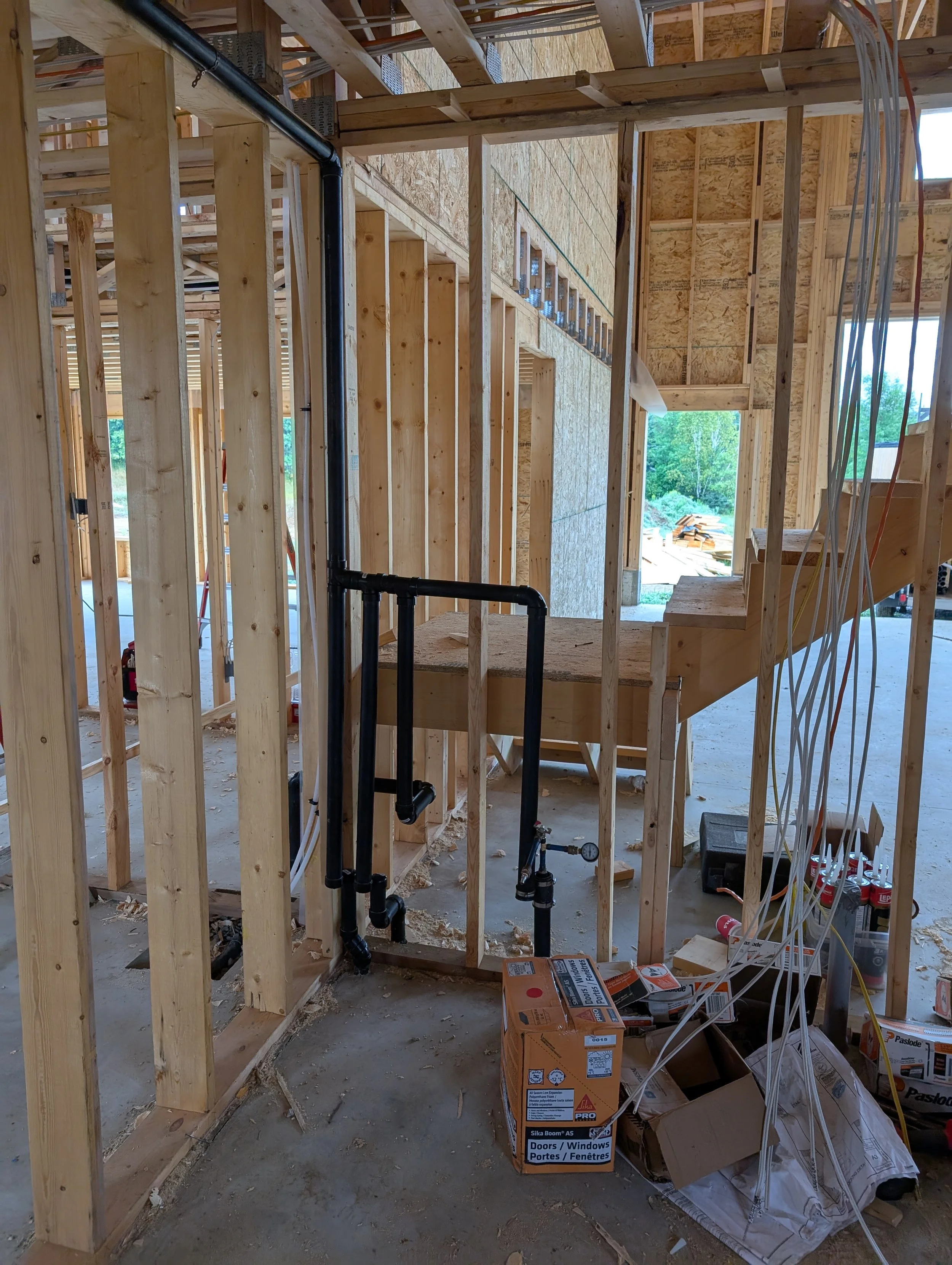 Interior view of a house under construction with exposed wooden framing, electrical wiring, and building supplies such as boxes of doors and windows.