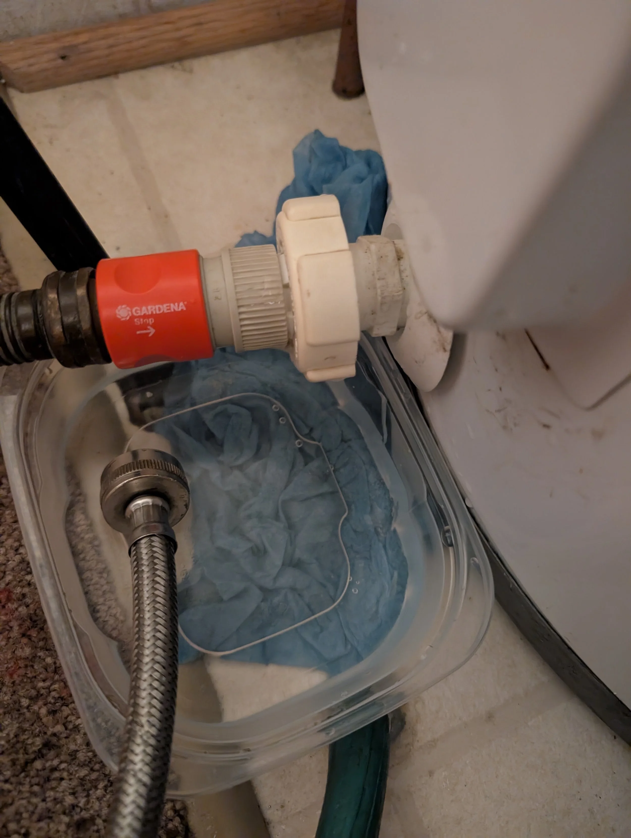 Close-up of a plumbing connection on a toilet with a flexible metal hose and a red shut-off valve labeled 'Gardena Stop'. The underlying area has a blue cloth or rag and some tools nearby.