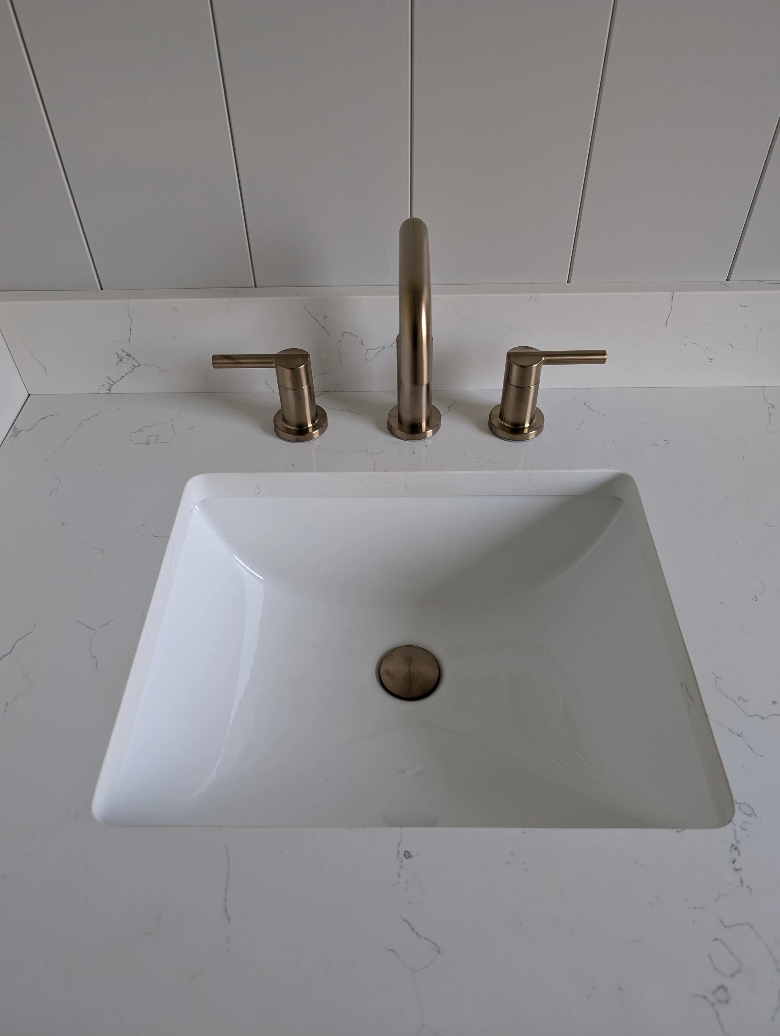 Brush nickel bathroom faucet with two handles mounted on a white marble countertop with grey veining, beneath a light grey, wood-paneled wall.