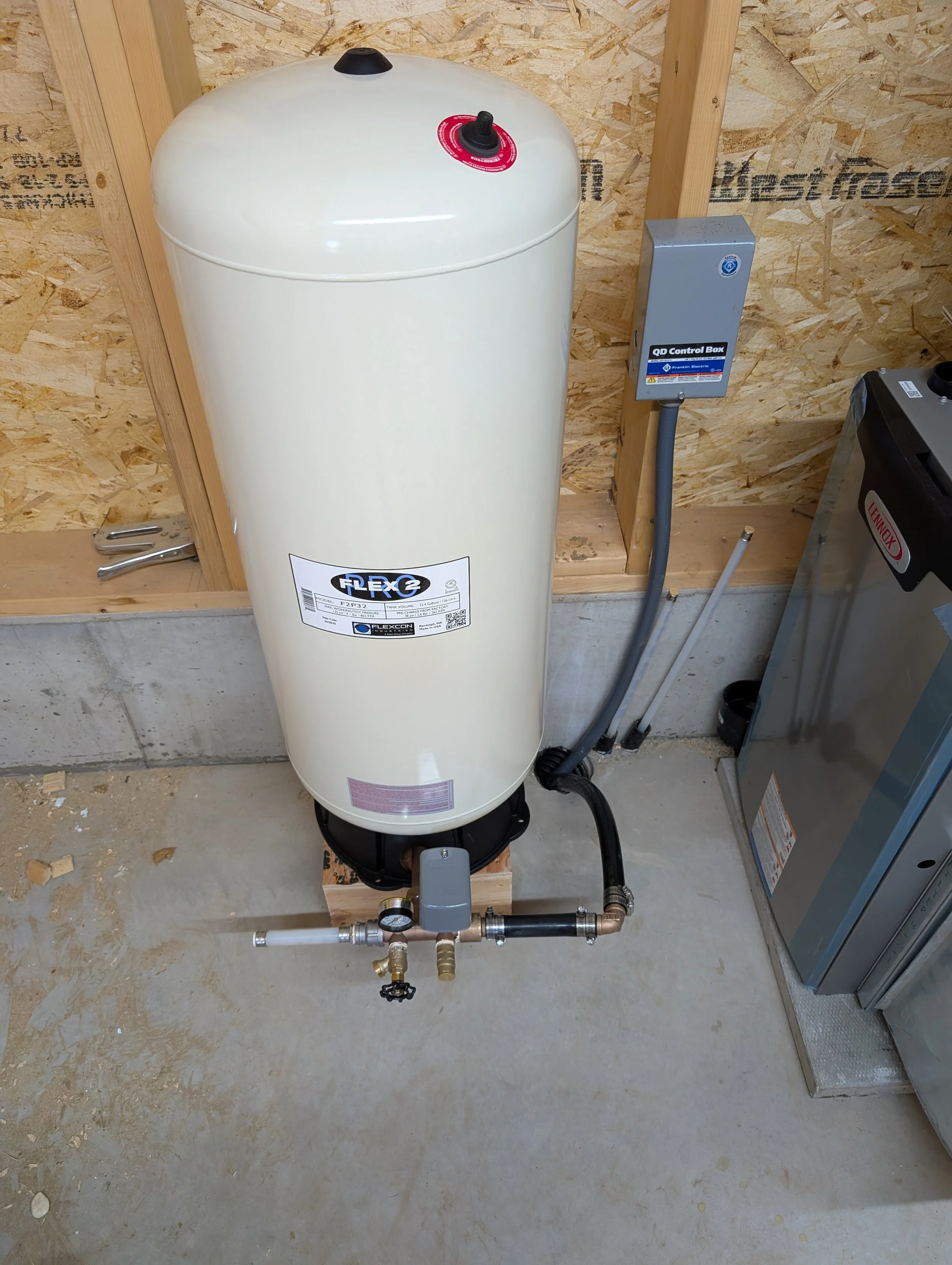 A large white flexcon water heater tank installed in a basement or workshop space, connected to plumbing and electrical components, with a wooden wall background.
