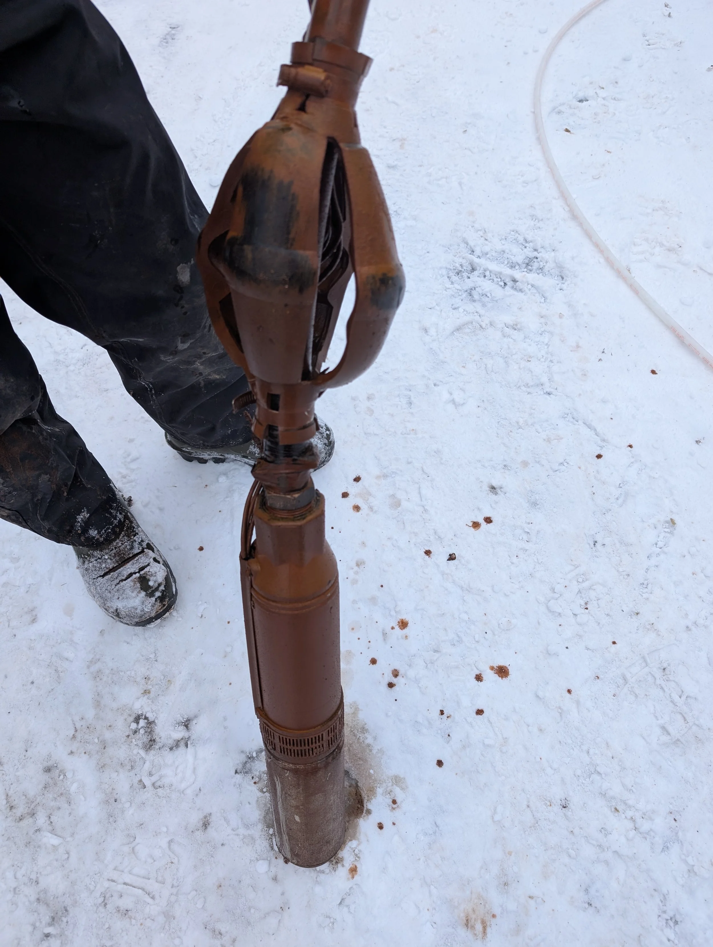 Close-up of a rusty pipe or mechanical device standing upright on snow-covered ground, with a person's black pants and shoes visible in the background.