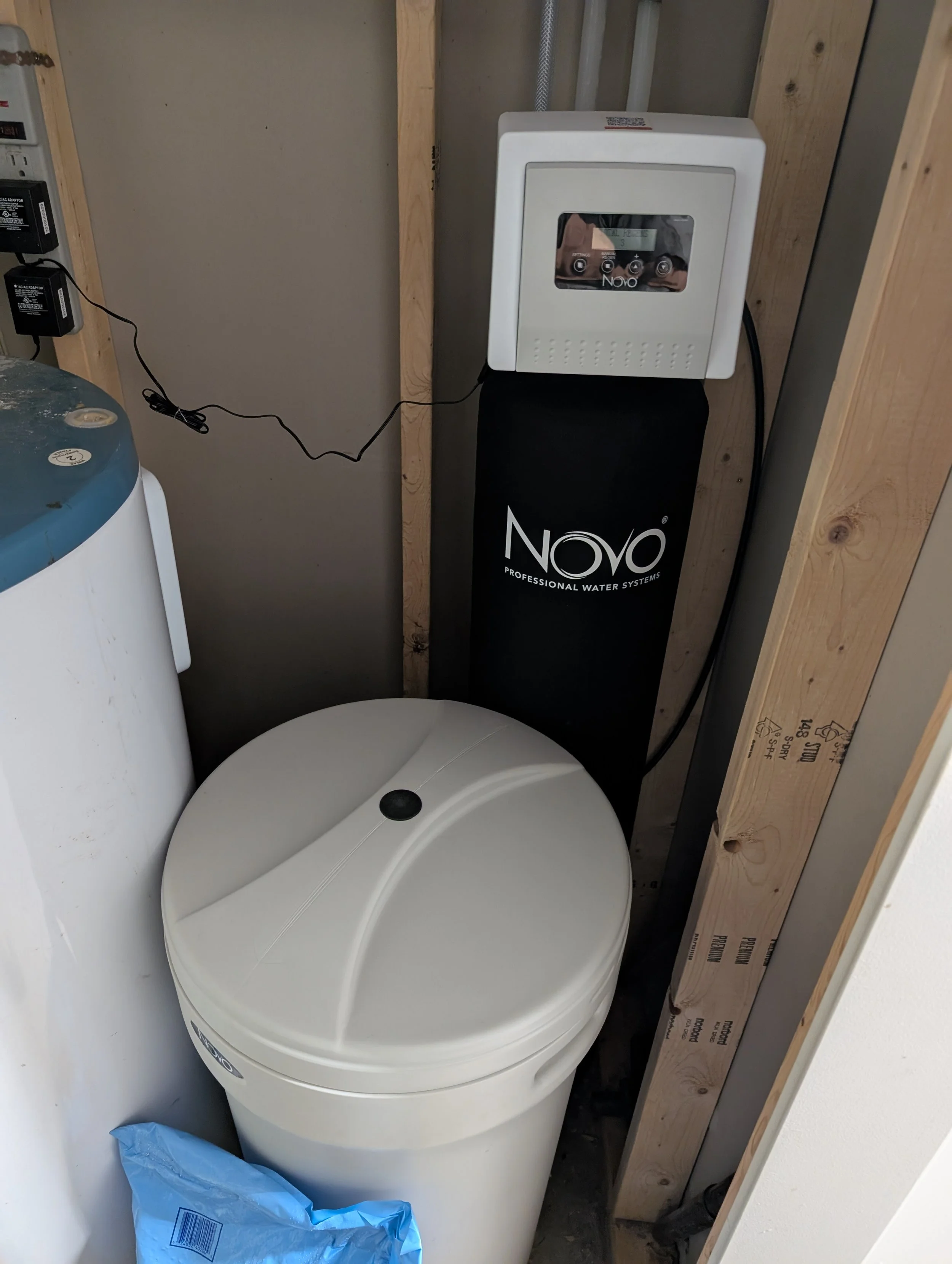 A water filtration system installed in a basement utility area with a white water filter, black water softener tank labeled 'Novo,' and a plumbing panel to the side. The area has unfinished wooden framing and exposed pipes.