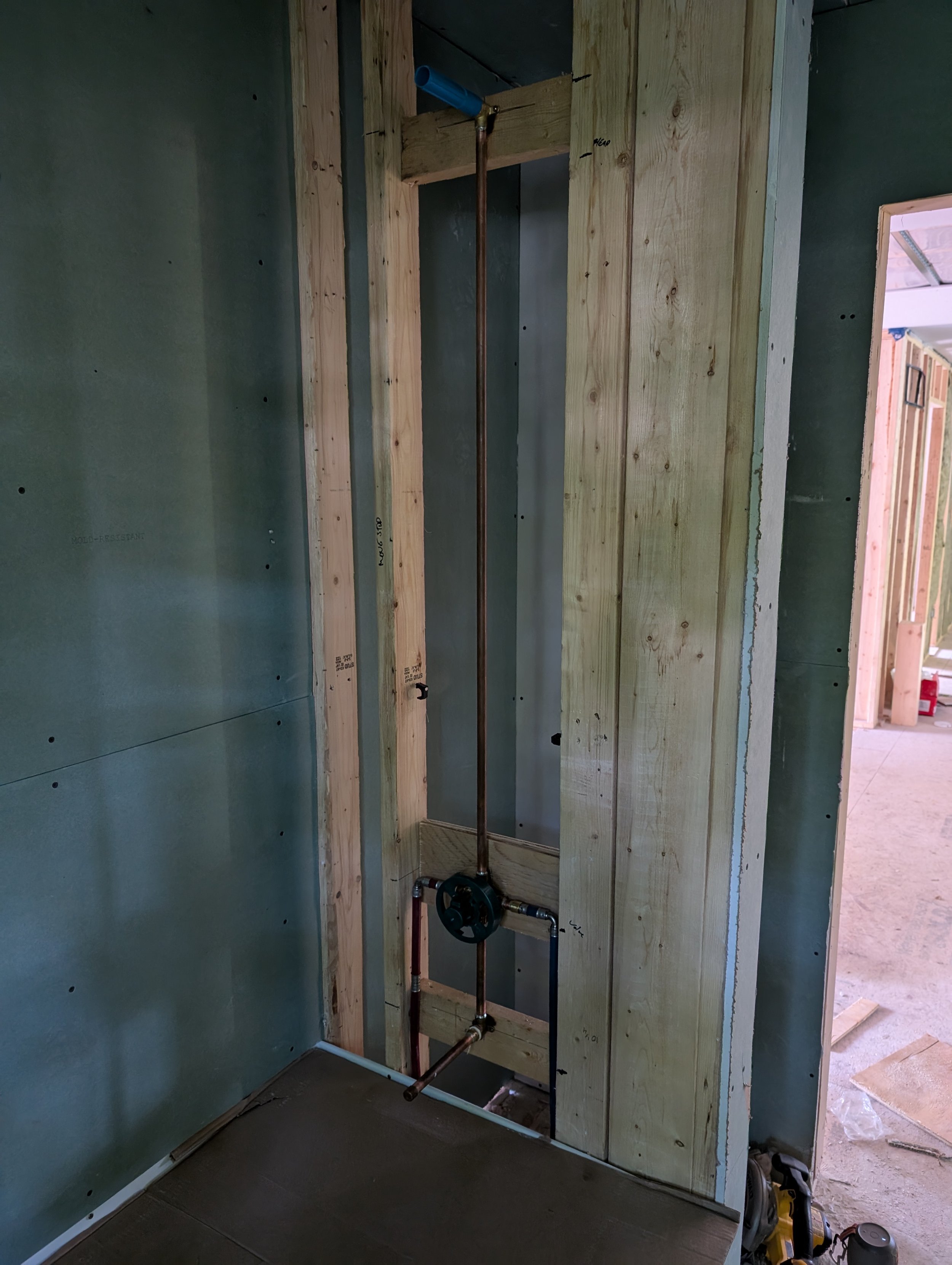 Plumbing rough-in in a building wall with copper and PVC pipes, a black anvil-style pipe adjustment tool, and a piece of drywall on the right side.