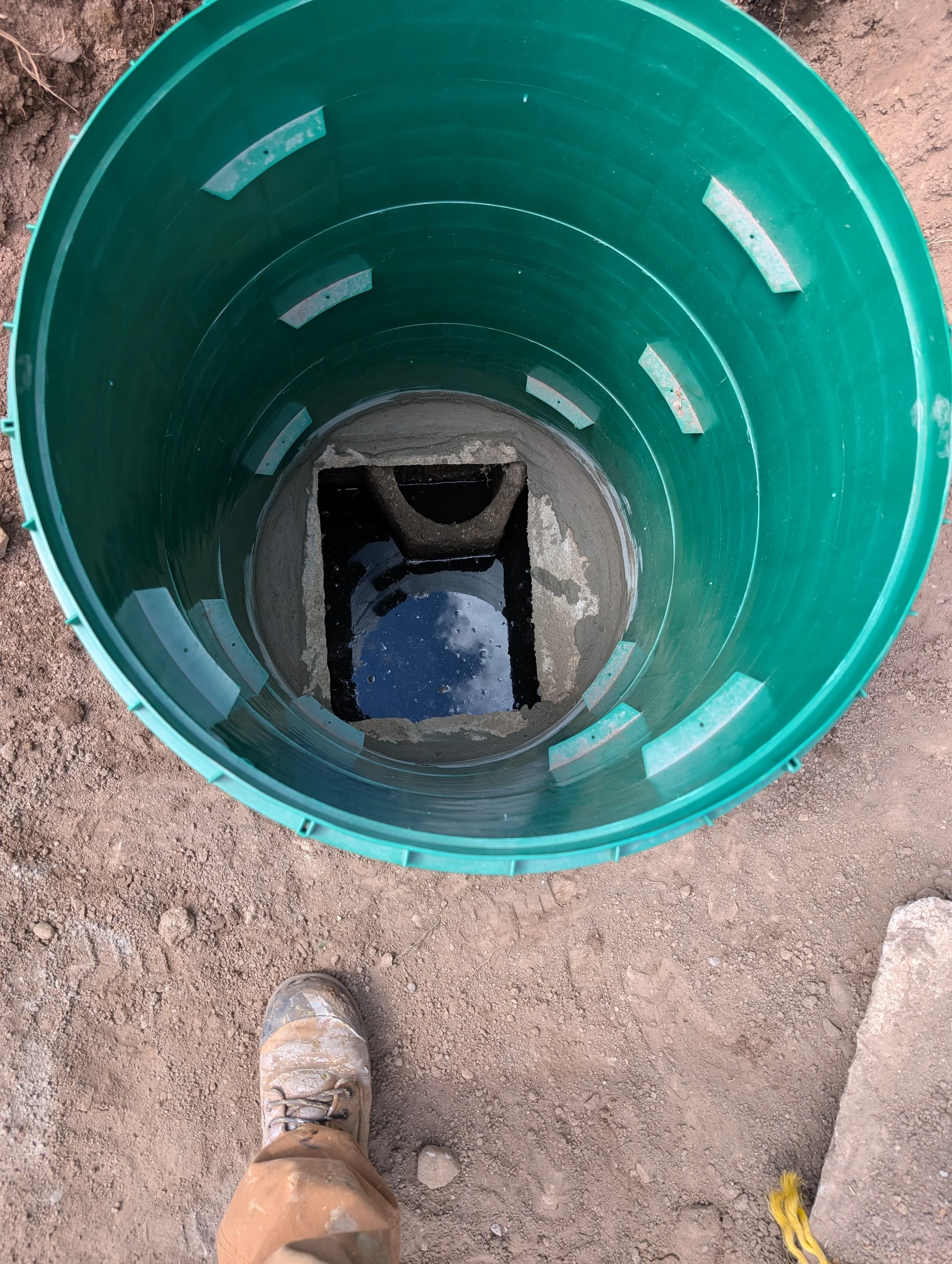 Looking down into a green cylindrical pipe with concrete and water at the bottom, on a dirt ground, with a person's worn hiking shoe and a yellow strap visible at the bottom of the image.