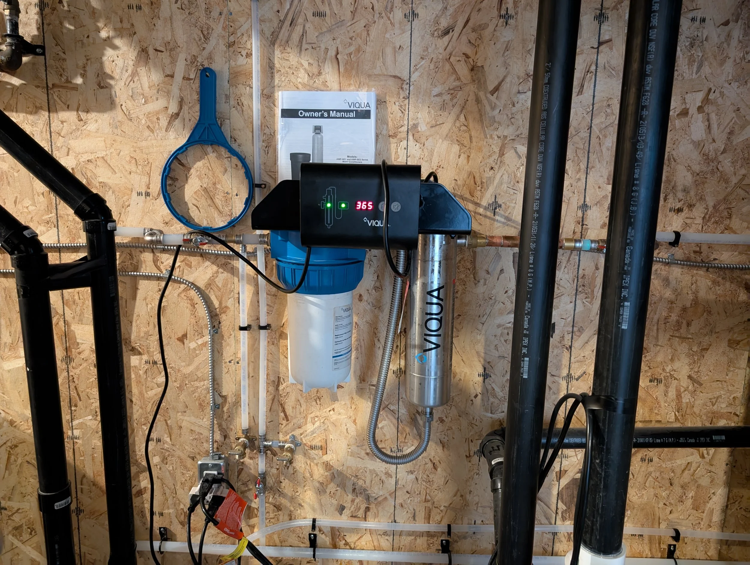 A plumbing system setup on a wooden wall with pipes, fittings, a water filter, and an owner’s manual, with a digital display showing the number 365.