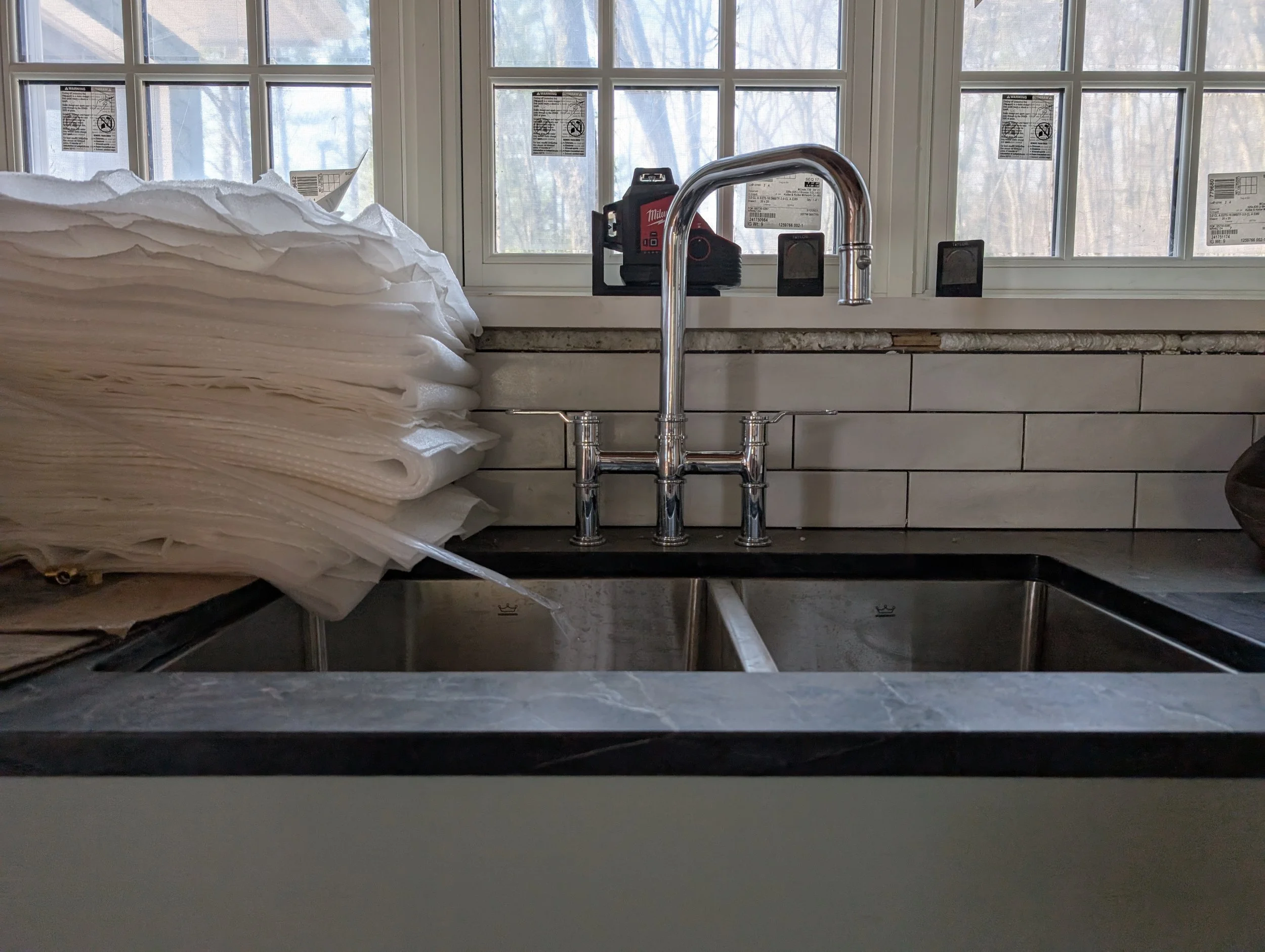 Kitchen sink with a chrome faucet, stacked paper towels on the left, and a window with multiple warning labels and stickers in the background.