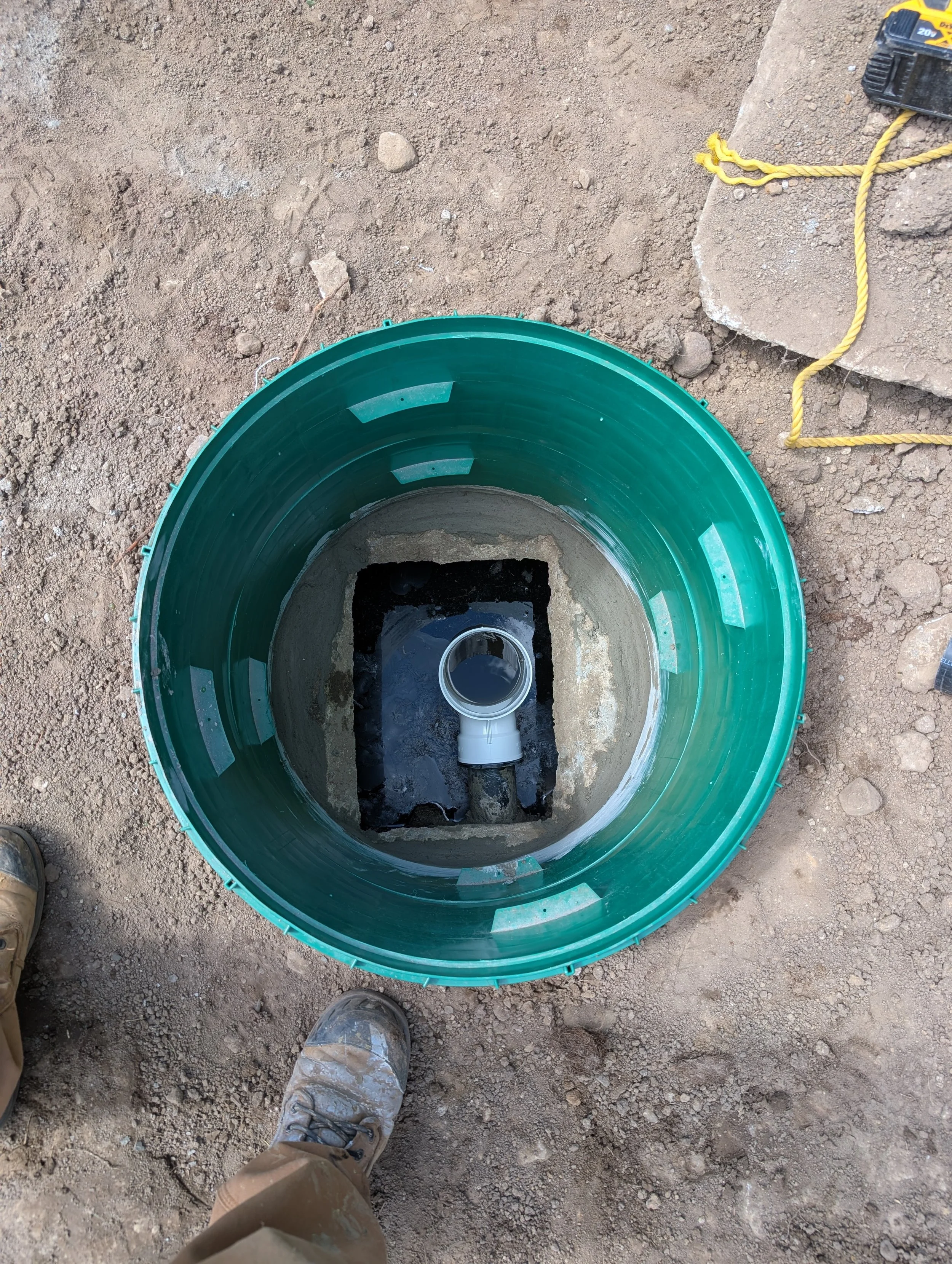 Construction worker standing on dirt ground, viewing a hole in the ground with a green circular cover and a pipe inside. Construction tools and yellow ropes are nearby.