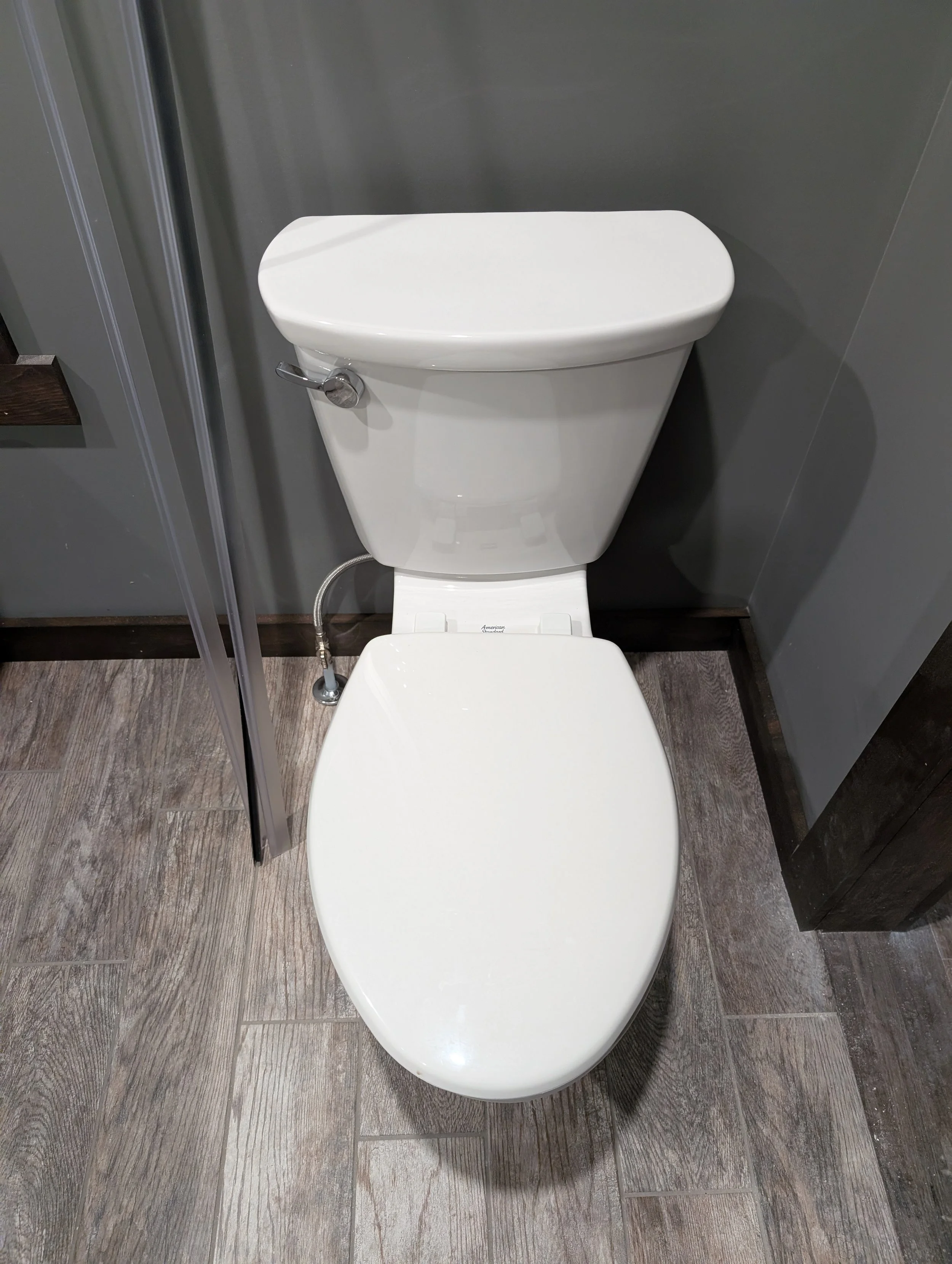 A white toilet with a closed lid in a modern bathroom with wood-look tile flooring and dark gray walls.