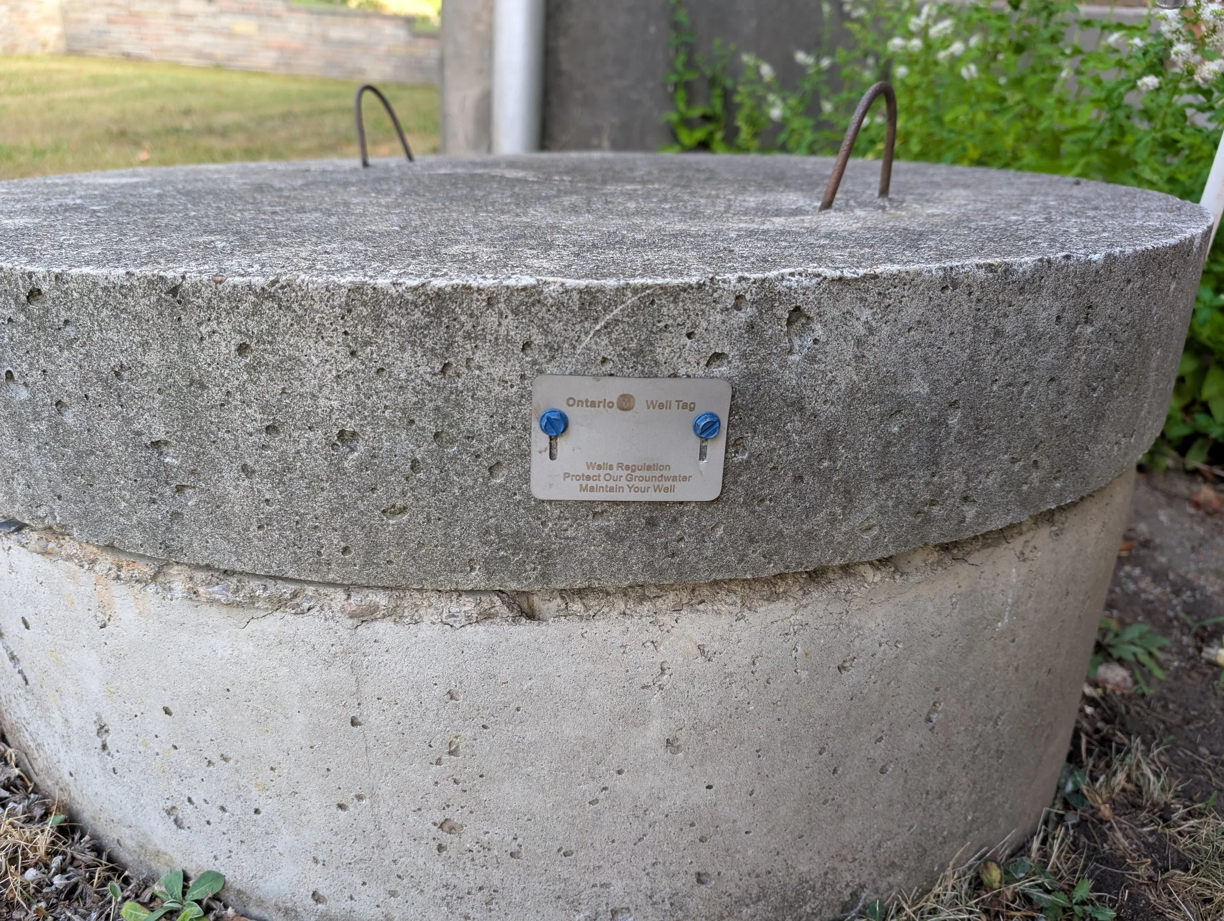 A large concrete well with metal handles on top and a metal cover, surrounded by grass and plants, with a small label indicating 'Wells Regulation'.