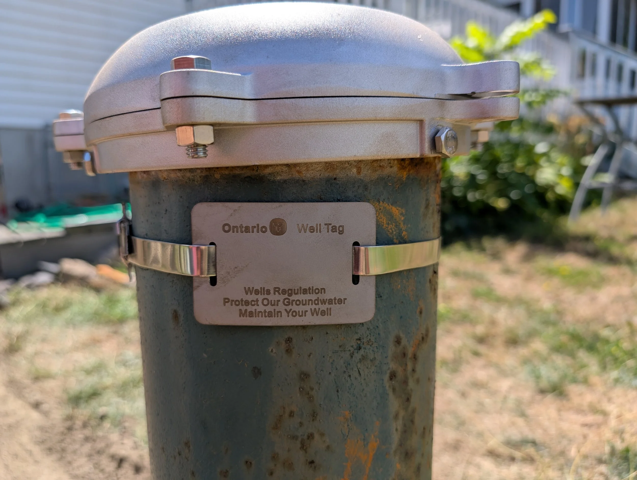 Close-up of a well tag on a ground water well, with part of the metal pipe and a background of grass and buildings