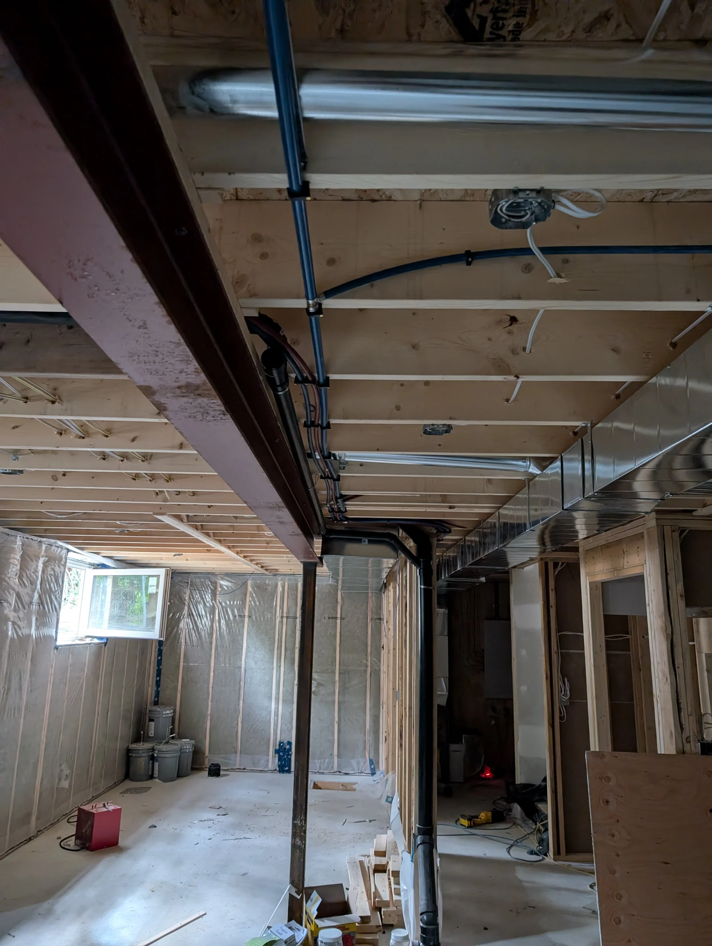 The photo shows an unfinished interior of a building under construction, with exposed wooden framing, plumbing pipes, electrical wiring, and construction materials scattered on the floor.