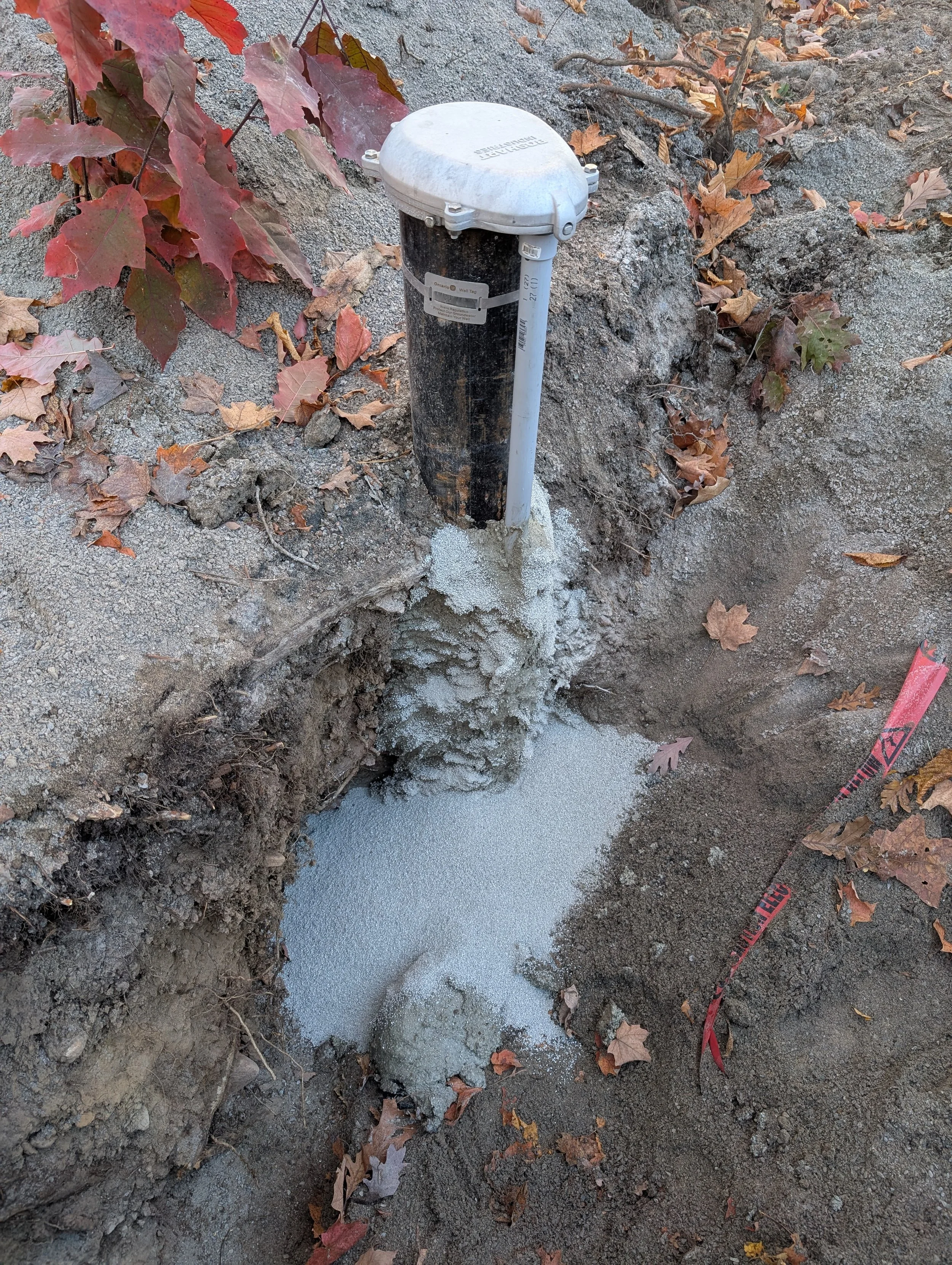 A buried water or utility valve box surrounded by dirt, fallen autumn leaves, and construction caution tape.