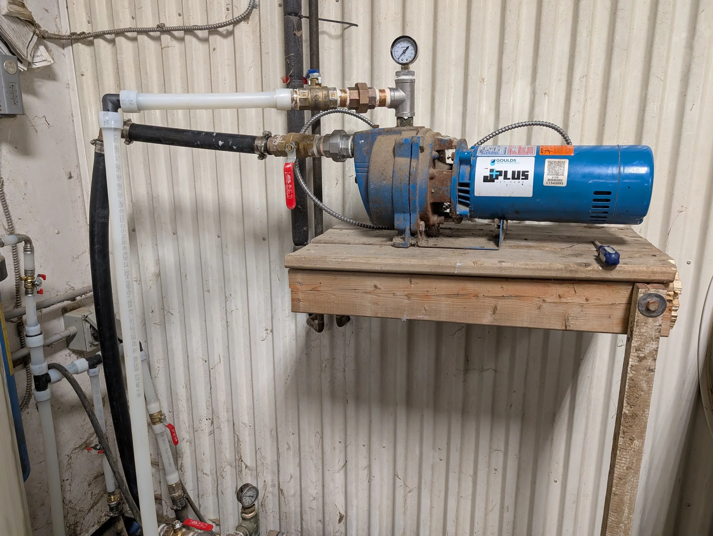 A blue Goulds jet pump connected to a series of pipes, gauges, and valves mounted on a wall in an industrial or utility setting.