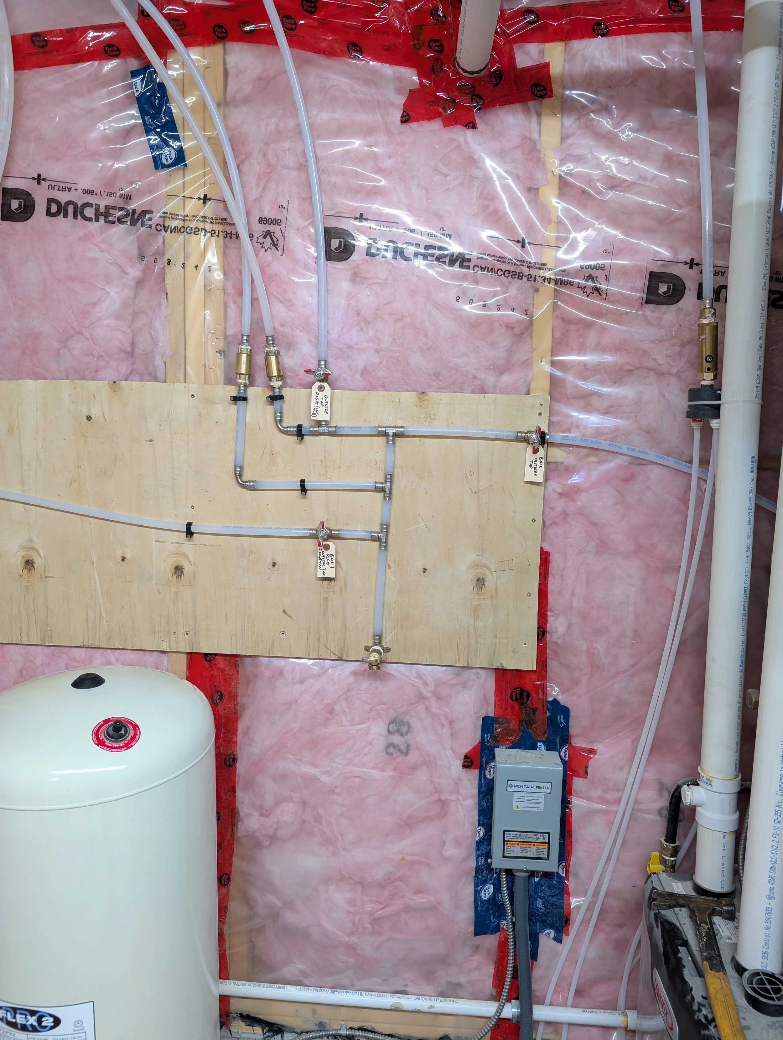 An unfinished plumbing installation in a wall with pink insulation, white pipes, a water heater on the left, a metal valve and a control box on the right, and various smaller pipes and fittings.